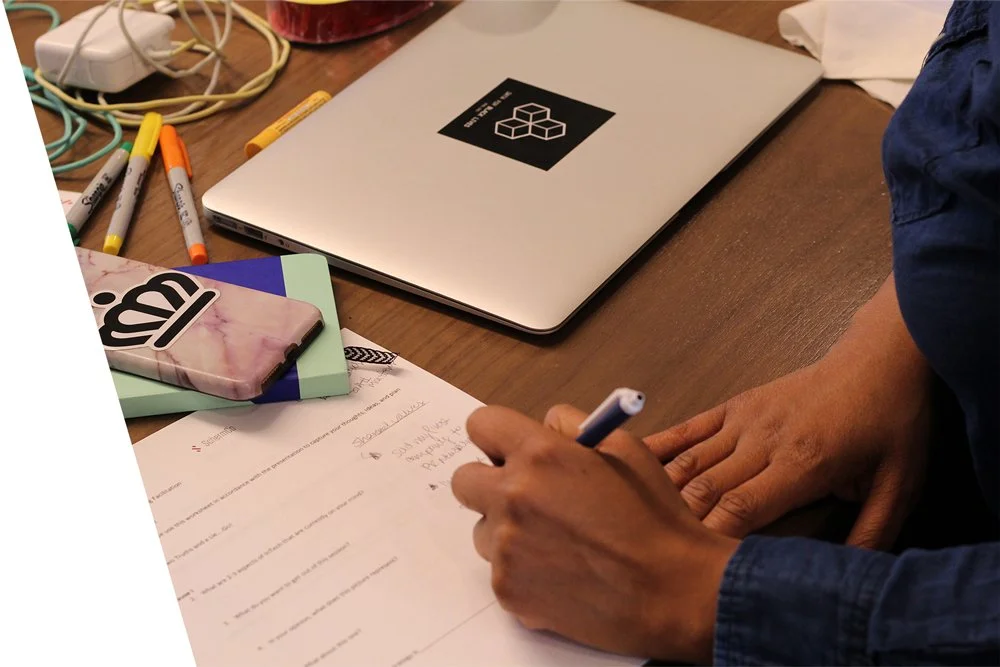 A wooden desk with a closed silver laptop, a smartphone with a pink and black case, several colored markers, a notebook, and a person's hands writing on a piece of paper.