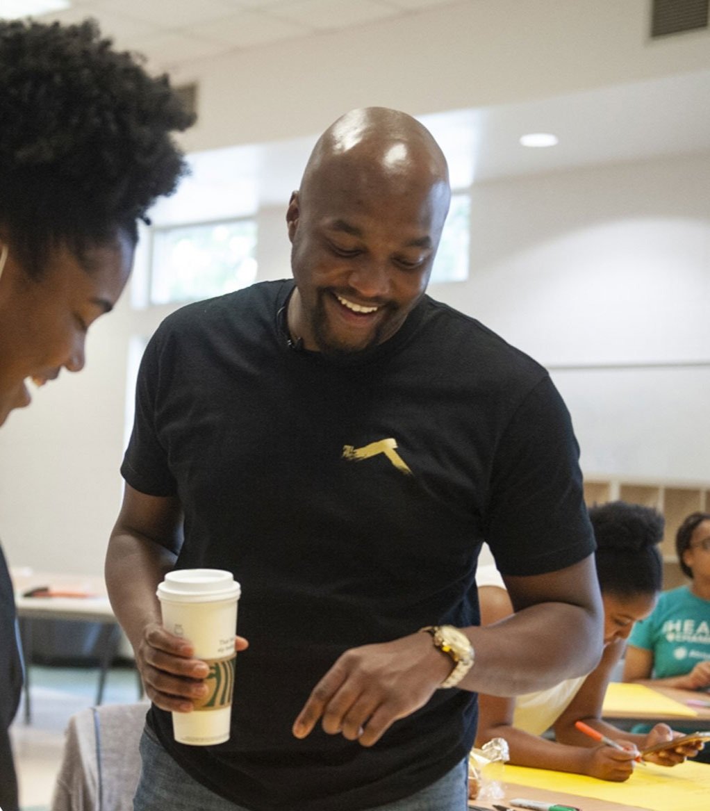A man smiling and holding a white disposable coffee cup, standing next to a woman in a classroom, with other students in the background.