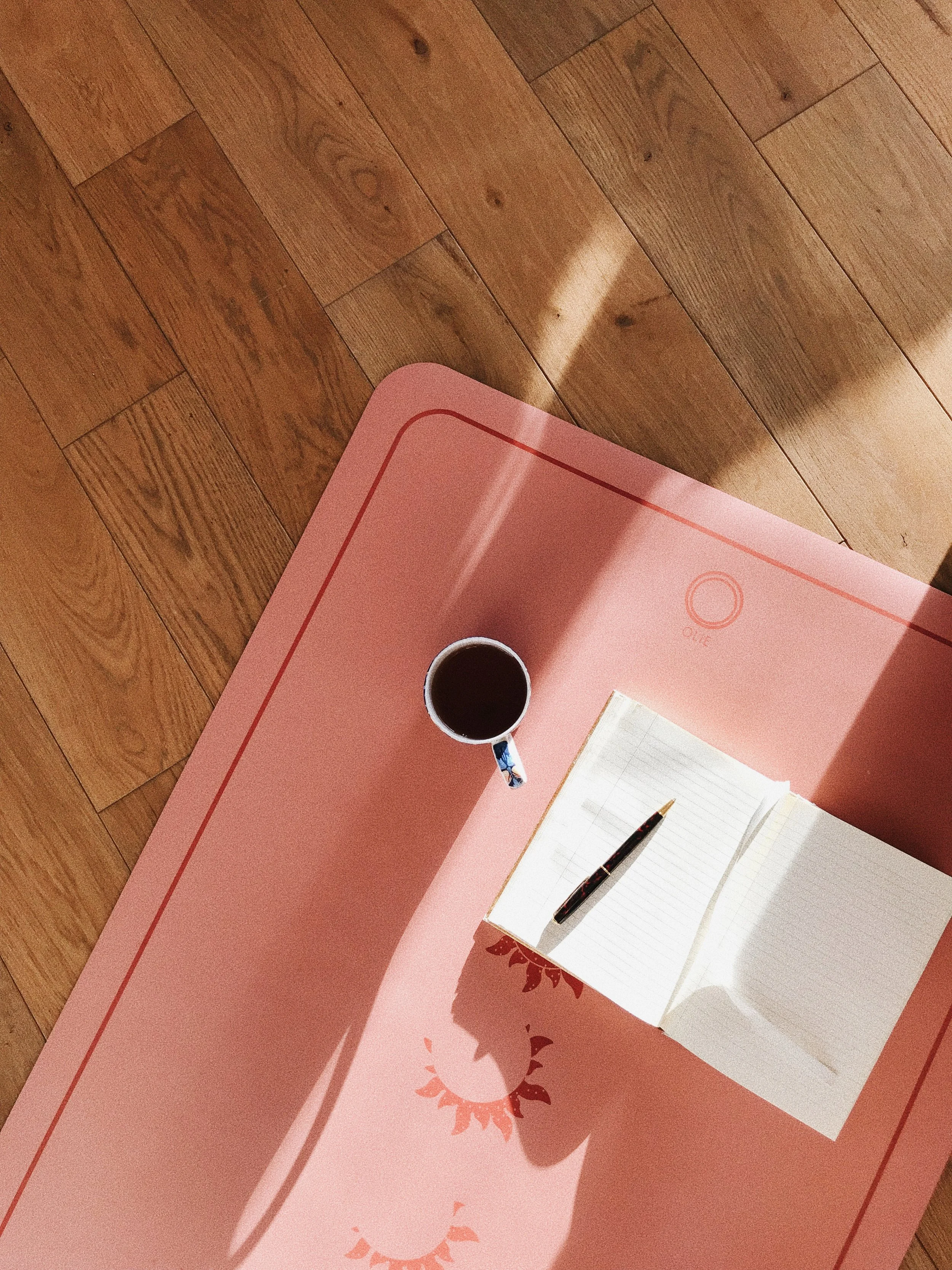 Open notebook and cup of coffee on pink yoga mat on wooden floor.