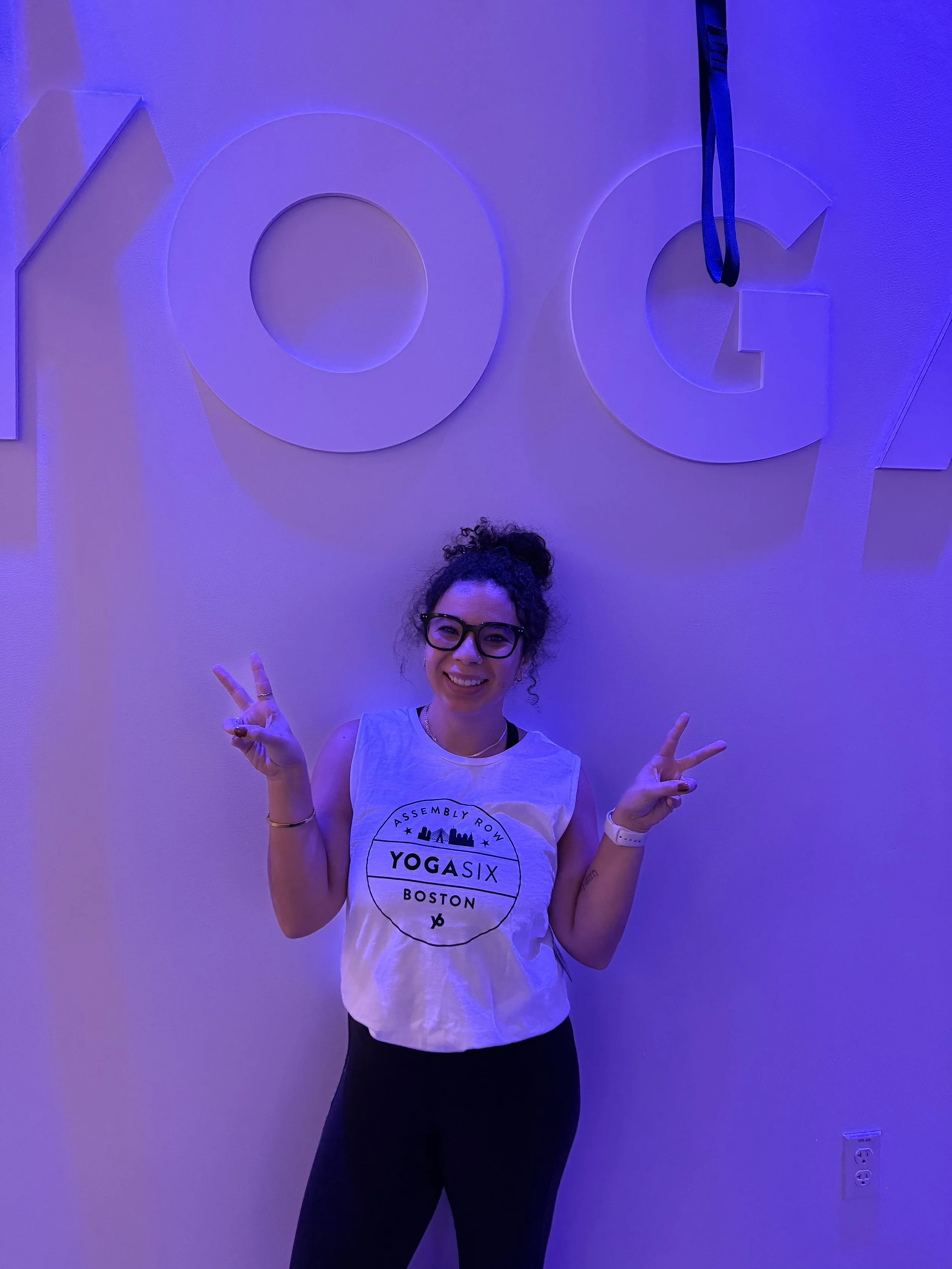 Woman with curly hair and glasses smiling and making peace signs in front of a wall with large white 3D letters for Google, under purple lighting.