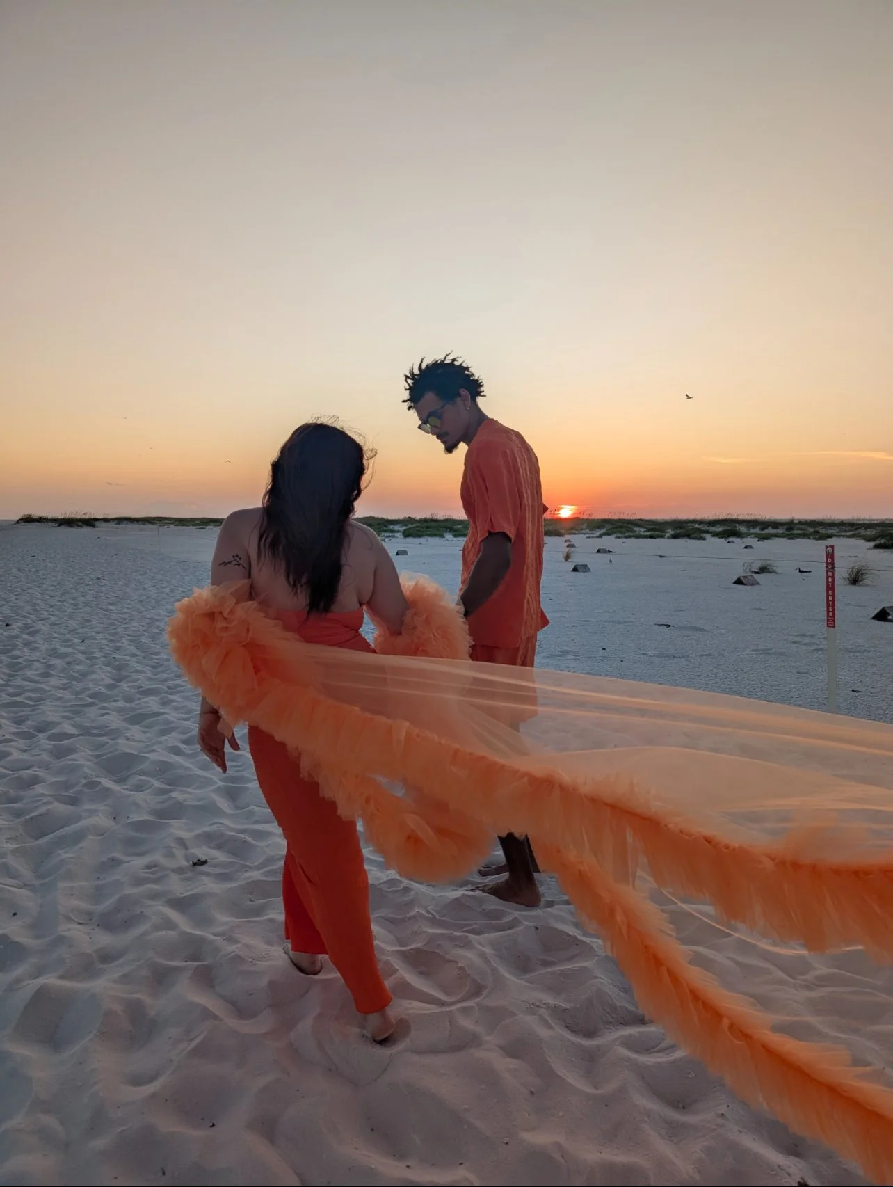 Two people in orange clothing walking on a sandy beach during sunset, with a long orange feathered fabric flowing between them in luxury attire.