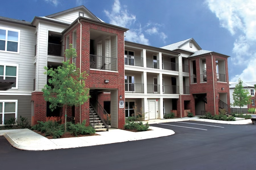 Modern multi-story residential building with red brick and white siding, featuring balconies, stairs, and parking spaces in the foreground, under a blue sky with some clouds.