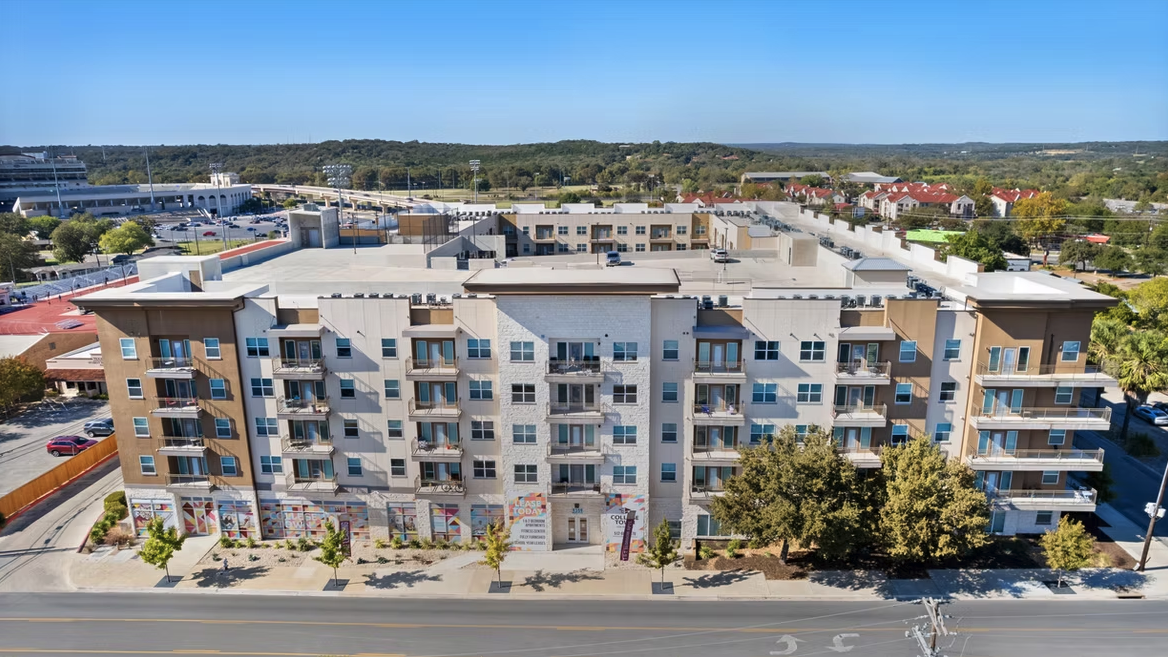 A multi-story residential building with commercial spaces on the ground floor, surrounded by trees and parking lot, with a clear blue sky and green hills in the background.