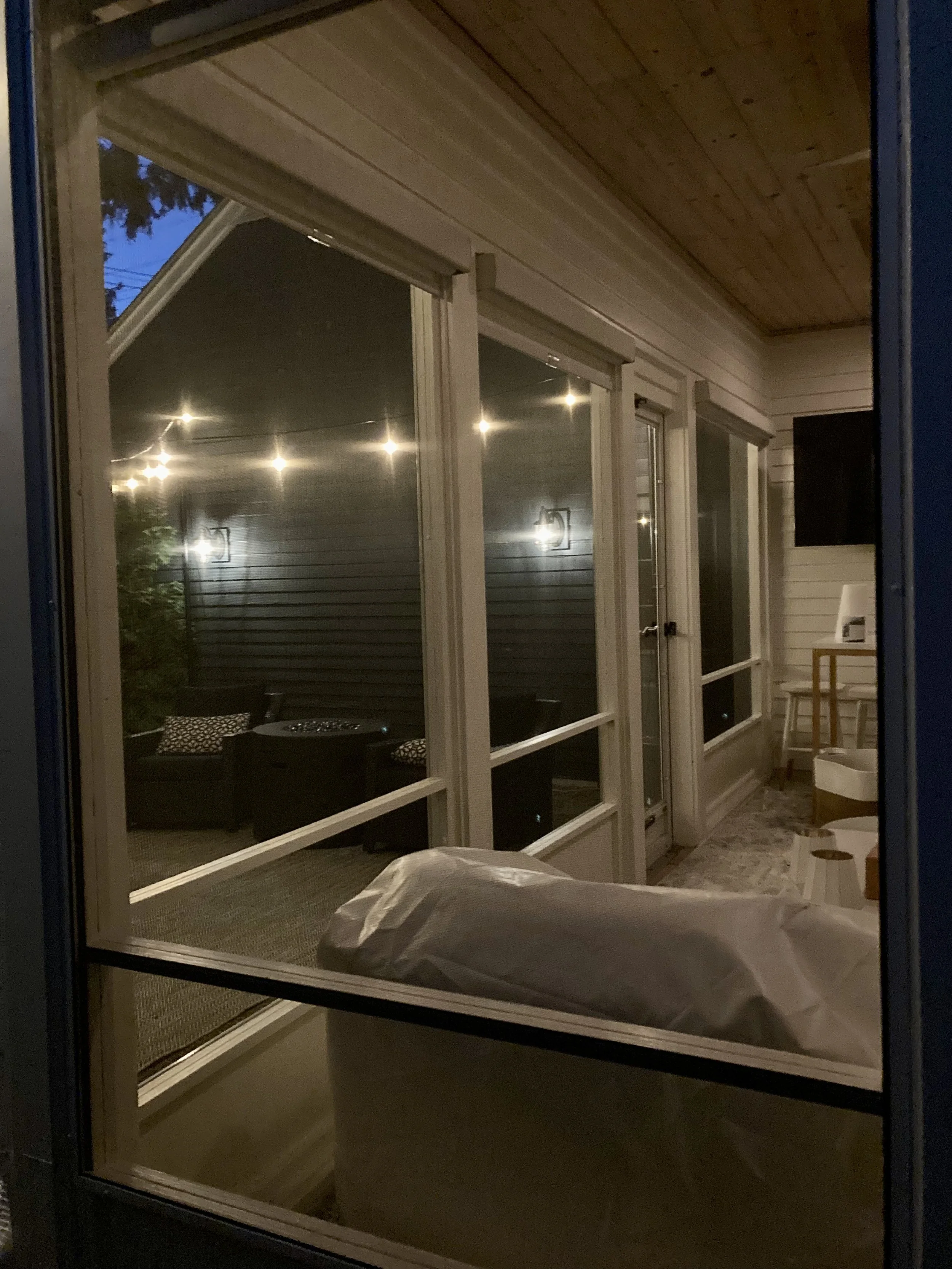 View through a glass door showing a backyard with string lights overhead, sitting area with chairs and a table, and part of a house with a wooden ceiling and siding.