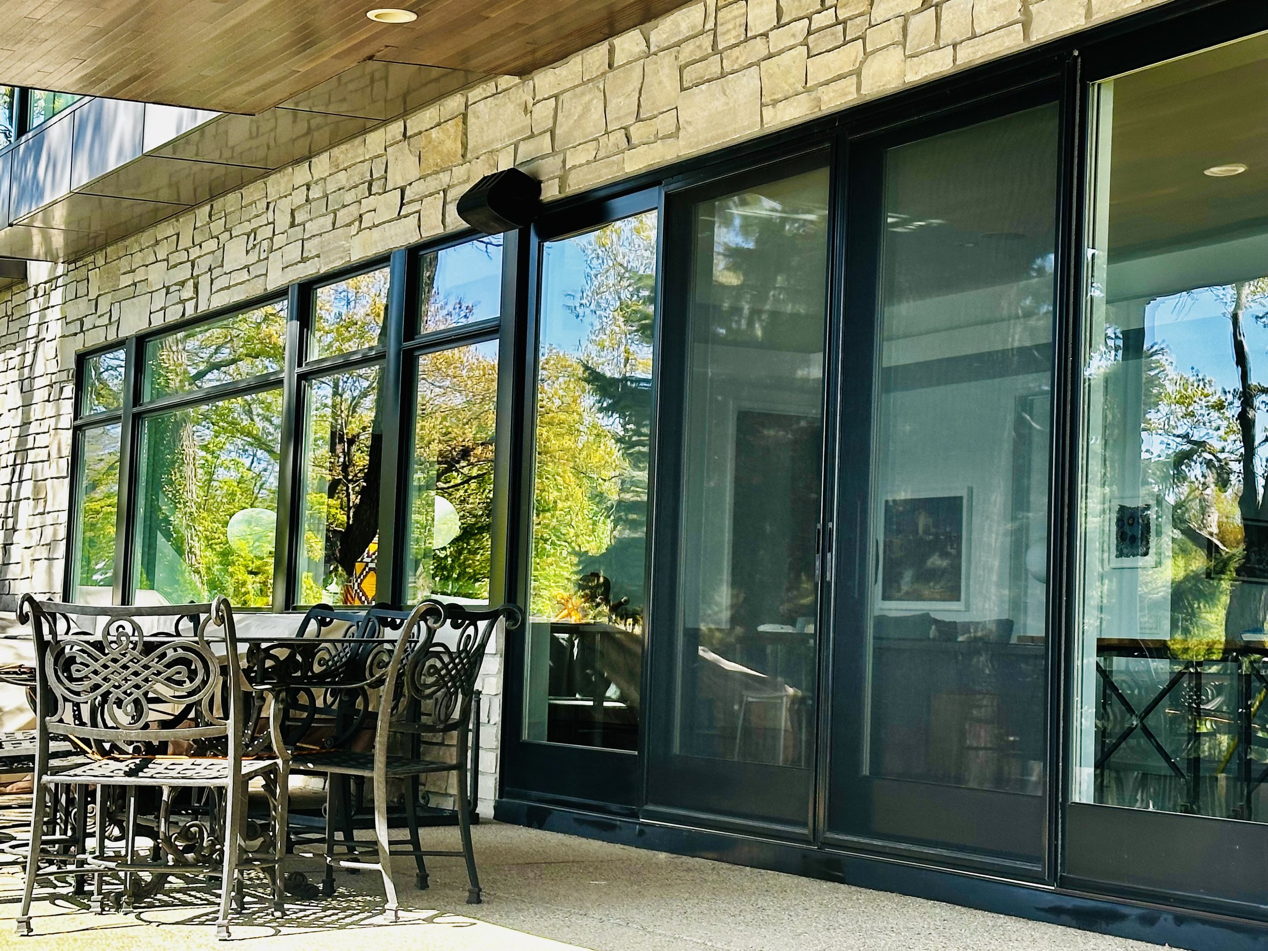 An outdoor patio with a metal dining table and ornate matching chairs in front of large glass sliding doors, with a stone wall and a tree reflected in the glass.