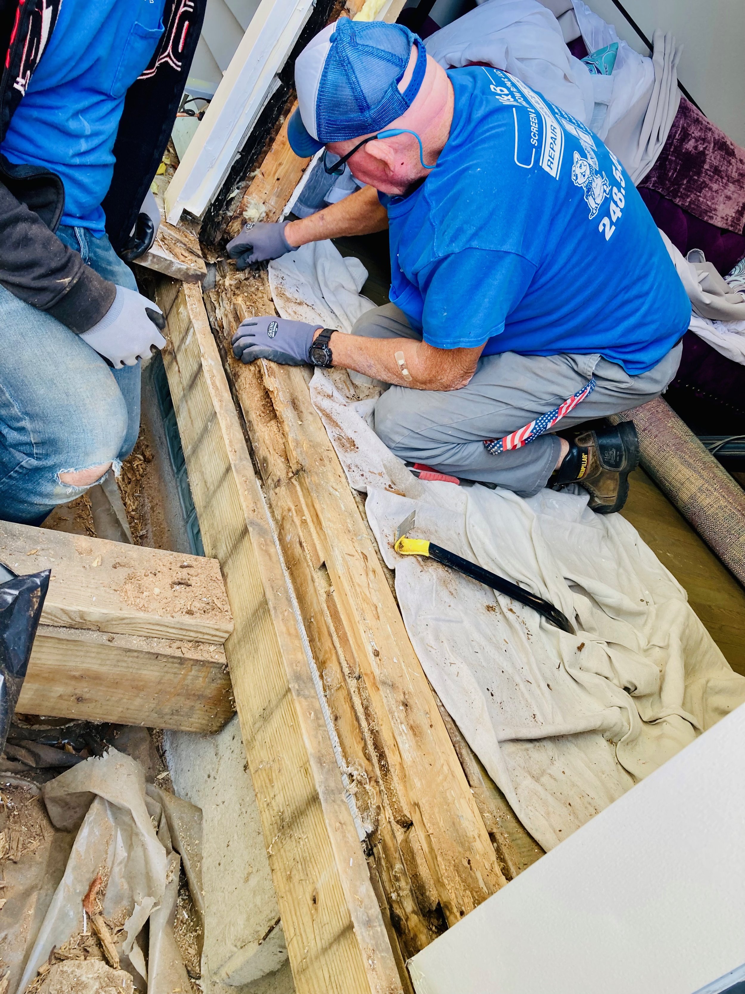 Man kneeling on a white cloth working on a wooden floor with a crowbar nearby and other tools, while wearing gloves, a blue shirt, a blue cap, and glasses.