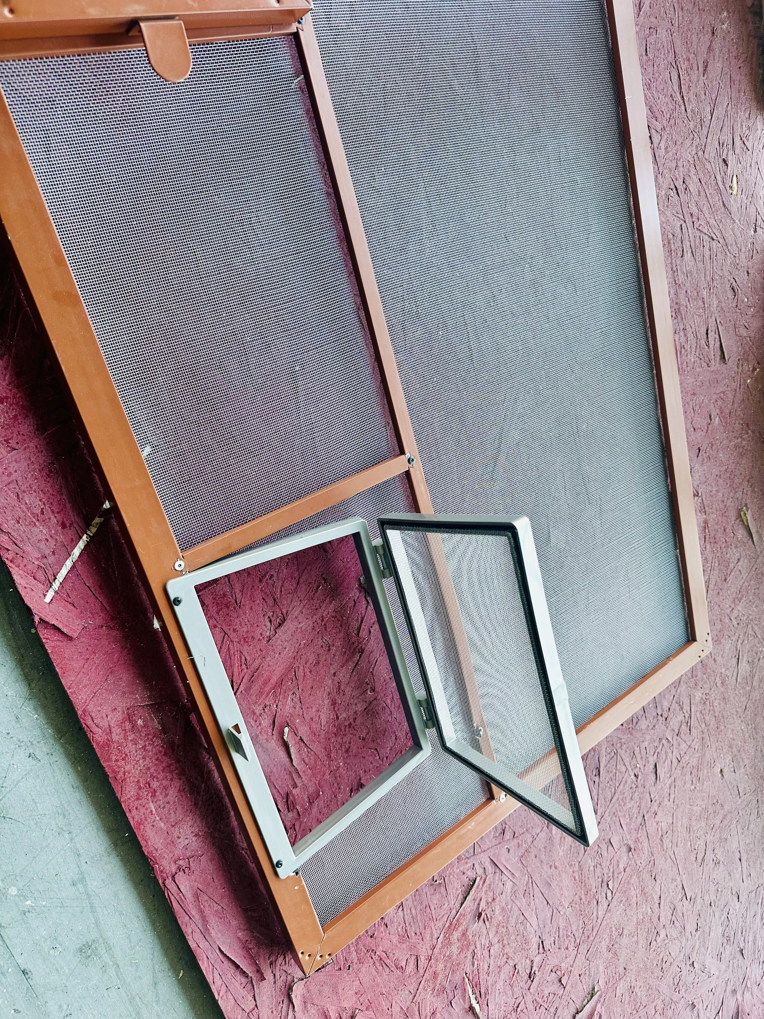 A rectangular wooden frame with mesh screening leaning on a pink wooden surface, with a partially open window frame in front of it.