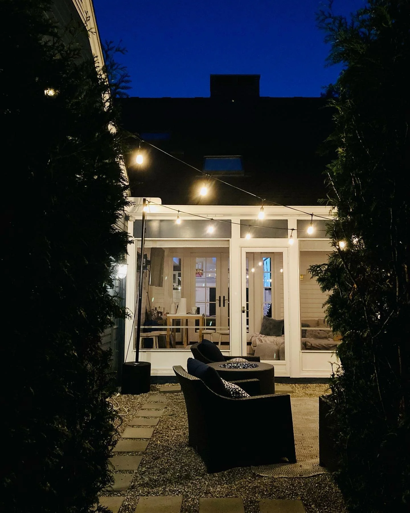 Nighttime outdoor patio with string lights, two black chairs with pillows, and a view into the well-lit interior of a house with glass doors and windows.