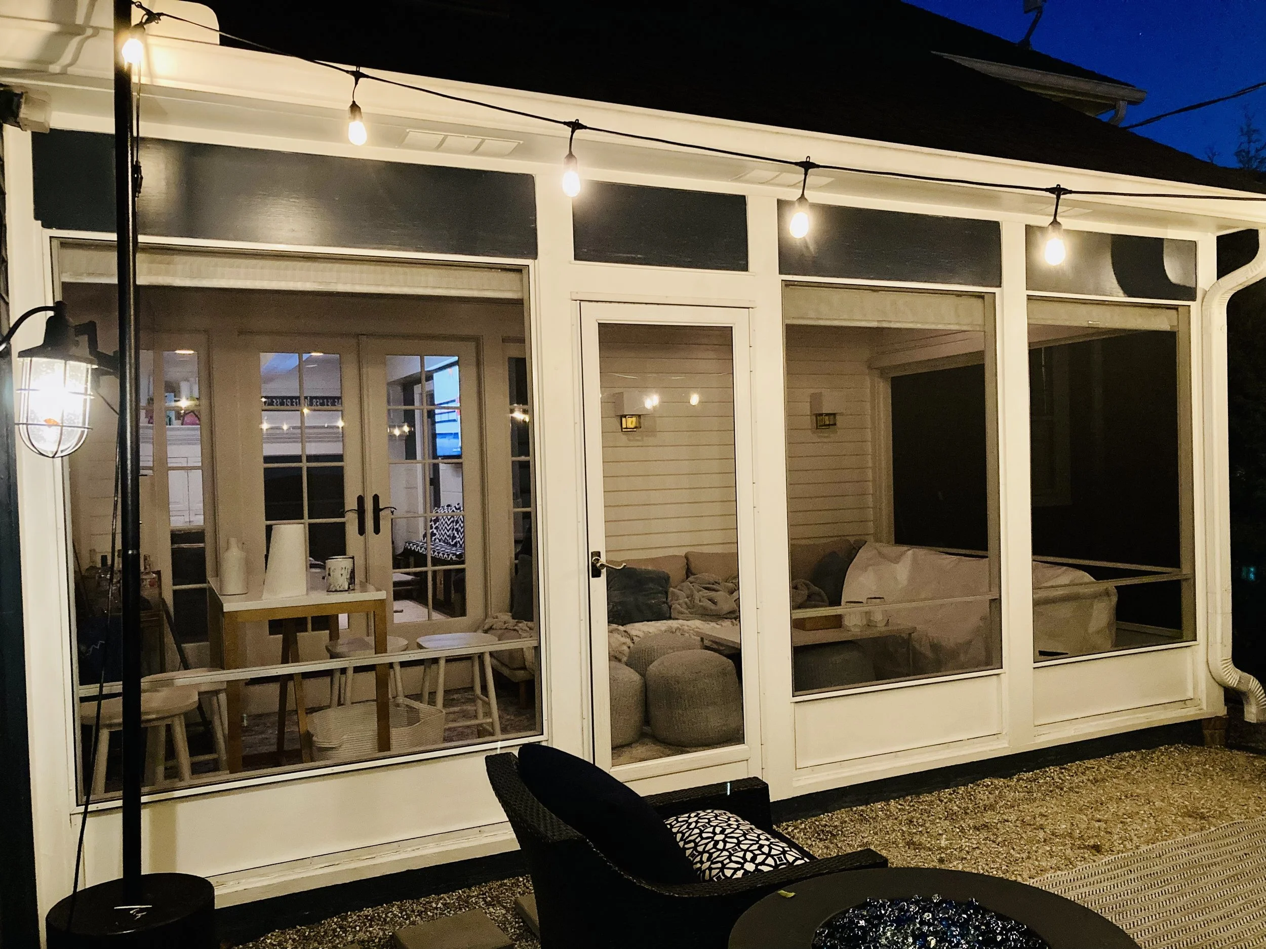 Nighttime view of a screened porch with string lights hanging across the top. Inside, there's a large gray sectional sofa, a table, and a dark-colored filing cabinet. The porch has glass doors leading into the house, and part of an outdoor chair with