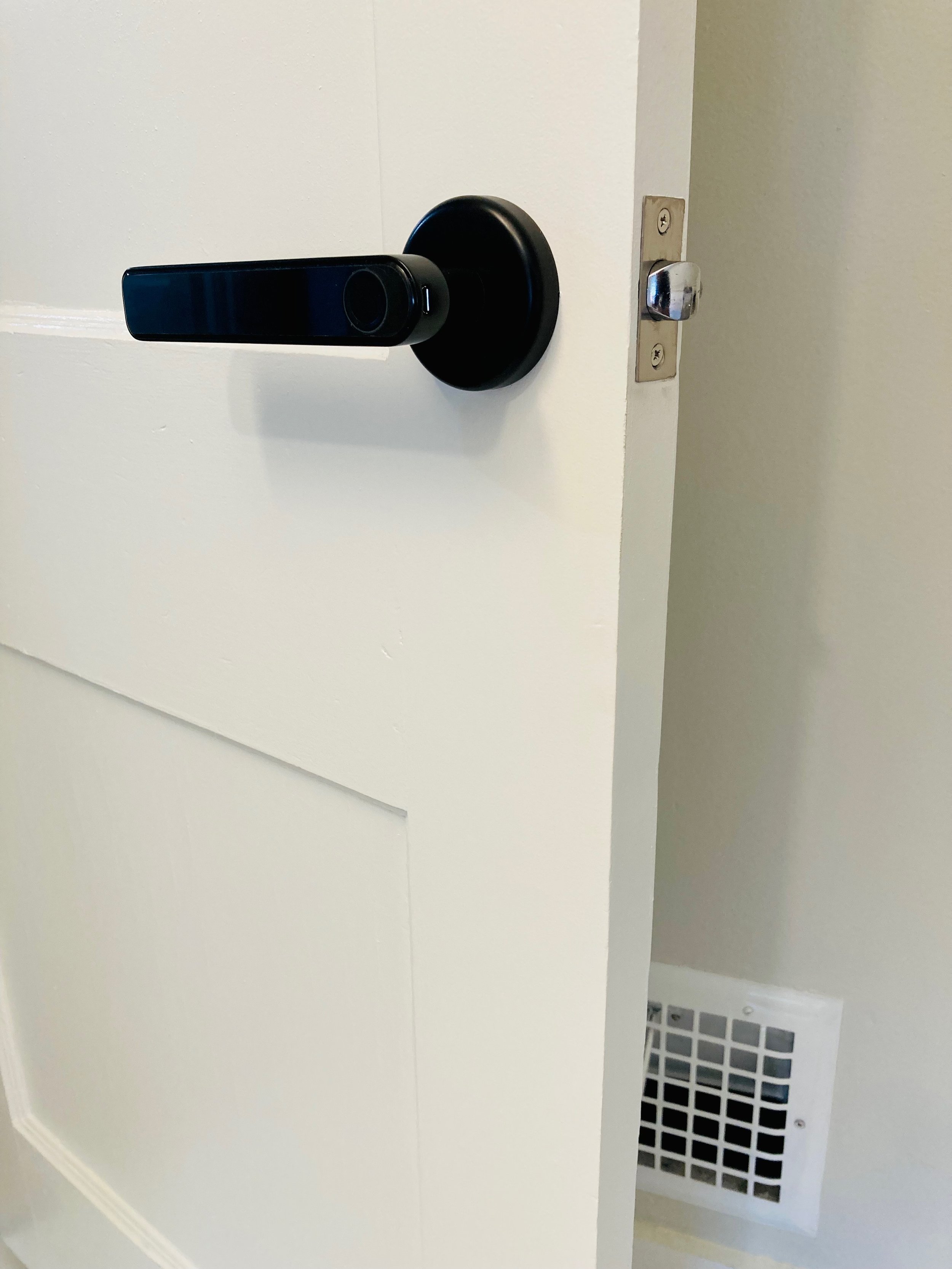 Close-up of a white door with a black digital door lock and a silver latch. Part of a white wall and a vent are visible in the background.