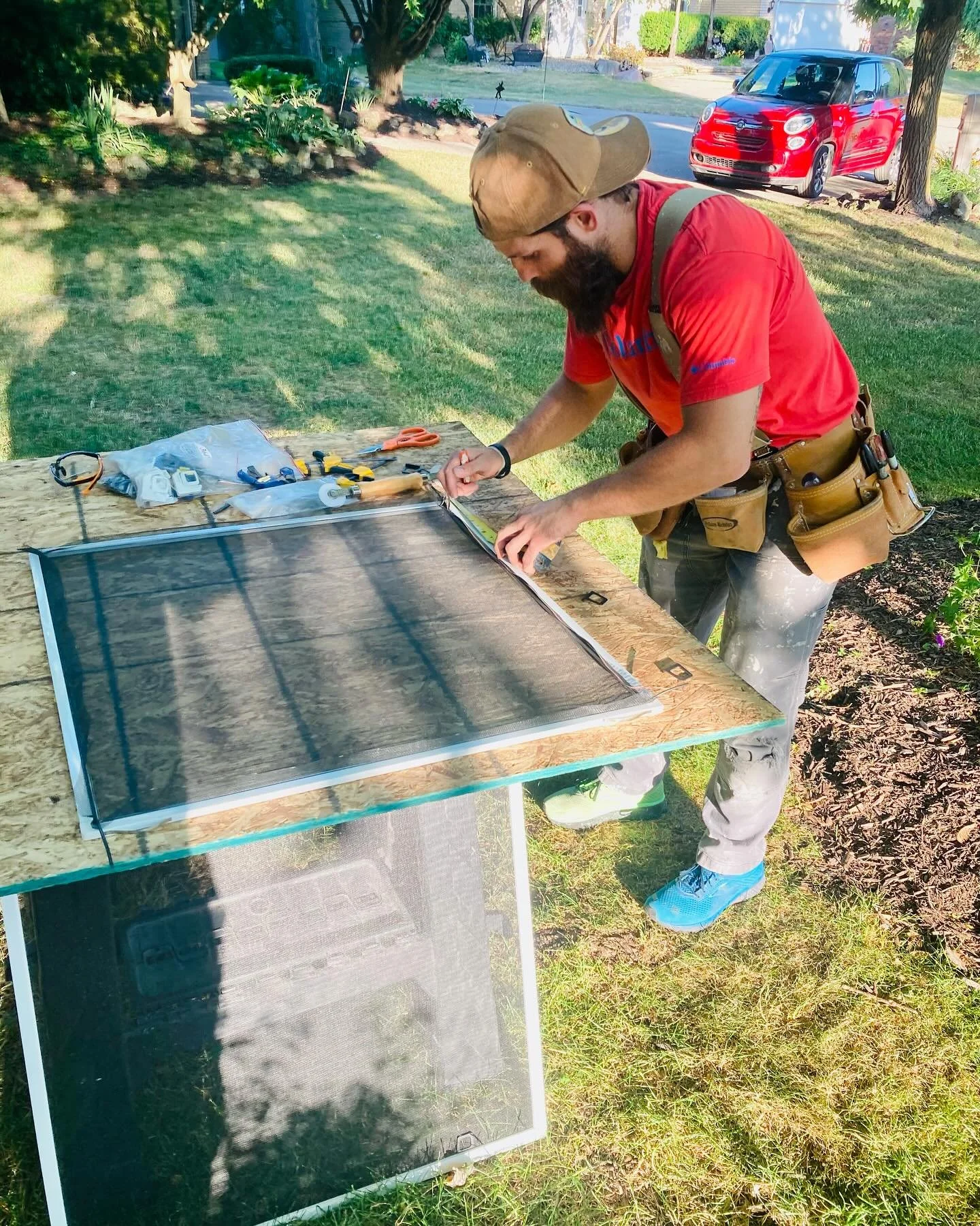 A man with a beard and a backpack installing a solar screen on a wooden work table outdoors in a grassy backyard with trees and a parked red car in the background.