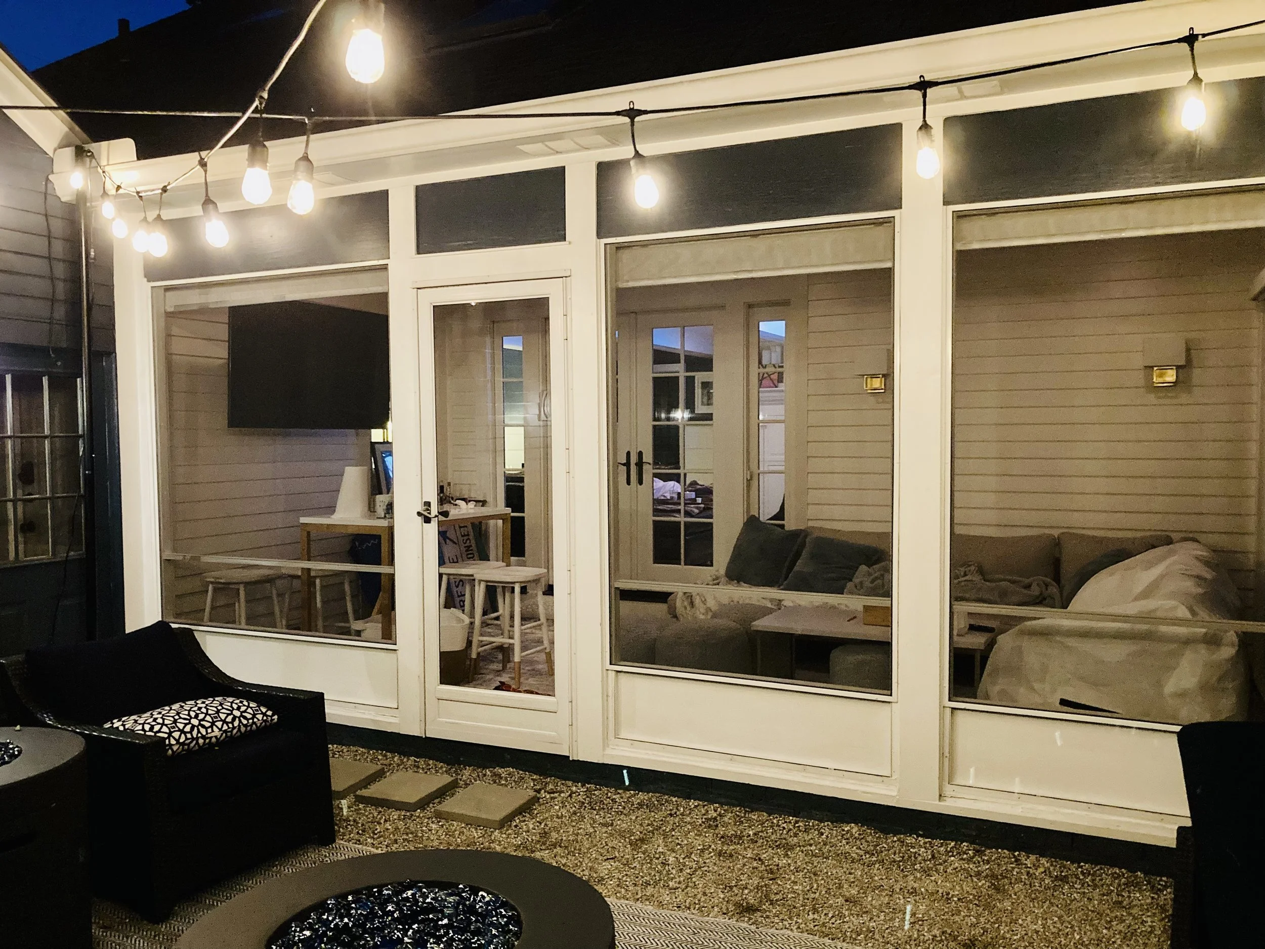 A backyard patio area at dusk featuring string lights hanging above a glass-enclosed room, with an outdoor seating area including a black sofa, a patterned cushion, and a round fire pit, along with a small table with stools inside the enclosed space.