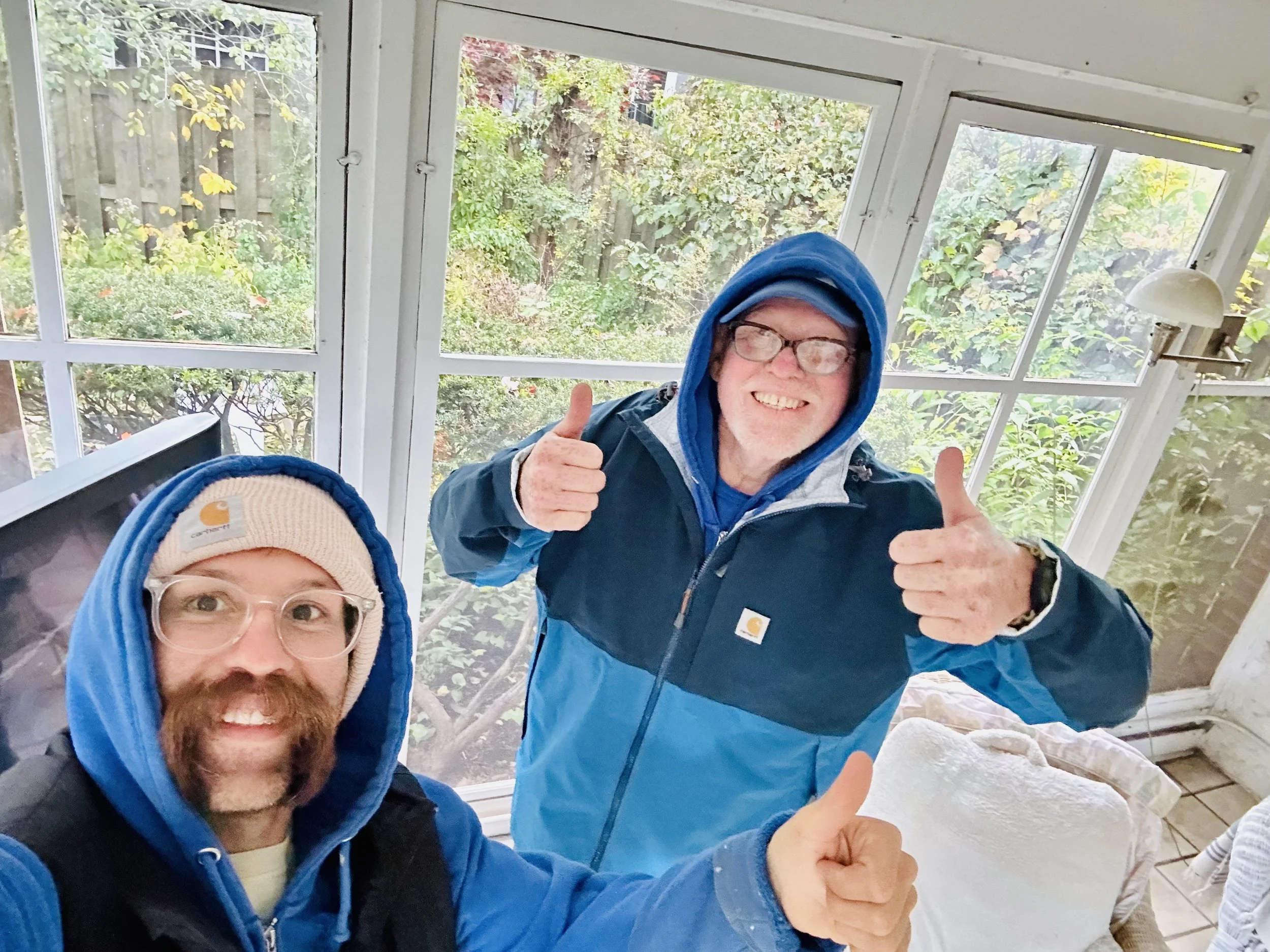 Two men smiling and giving thumbs-up in a sunroom with large windows and greenery outside, both wearing blue jackets with hoodies and beanies.