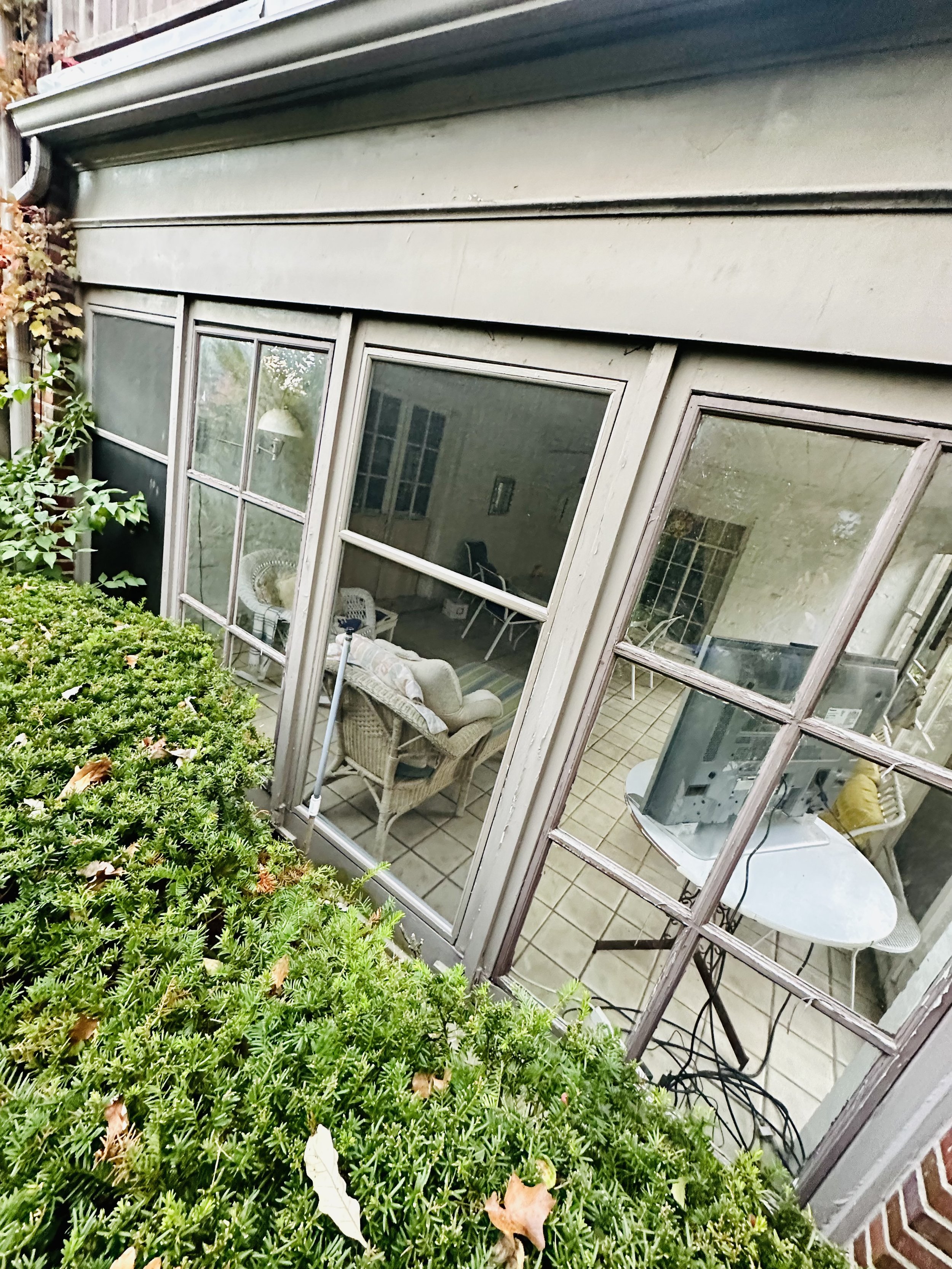 View of a screened porch from outside, showing green bushes and plants on the left, and the interior with chairs, a table, and various household items inside.