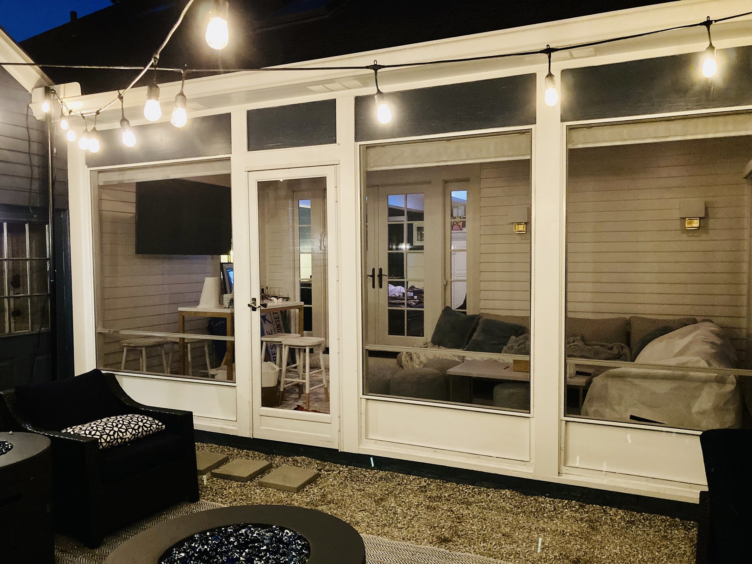 A backyard patio area at dusk featuring string lights hanging above a glass-enclosed room, with an outdoor seating area including a black sofa, a patterned cushion, and a round fire pit, along with a small table with stools inside the enclosed space.