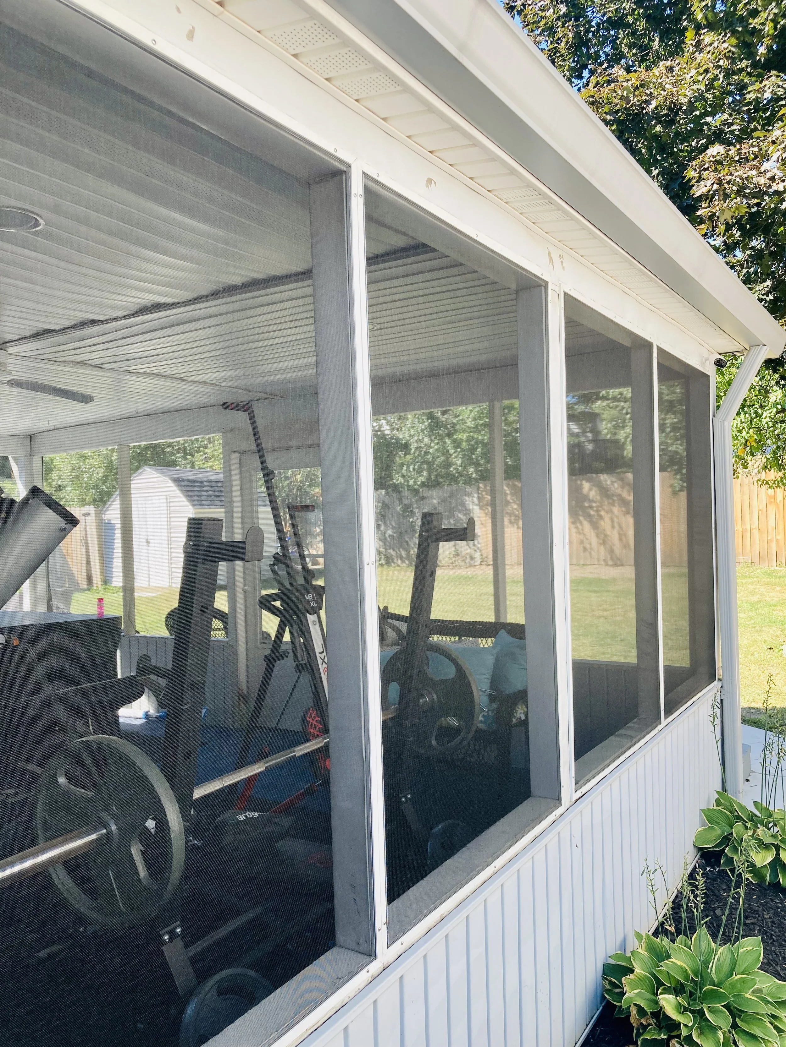 Inside a backyard screened porch with exercise equipment including a treadmill, stationary bike, and weights. The porch has white framed screens and a ceiling with a recessed light. Part of a garden with green plants is visible outside.