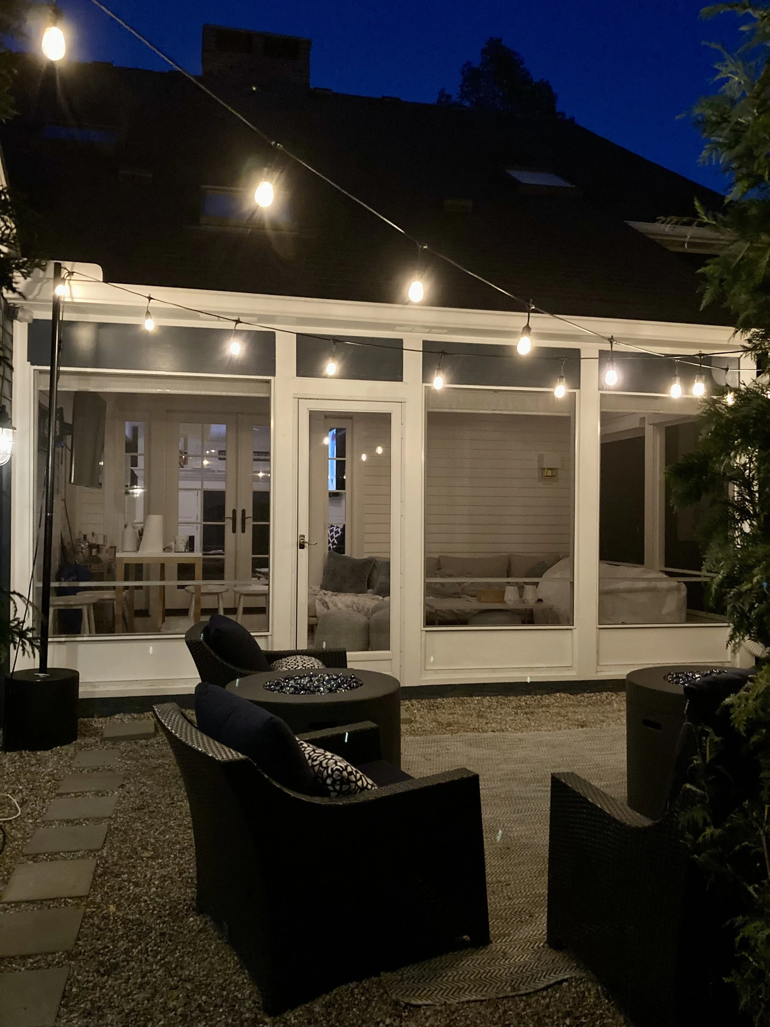 Nighttime outdoor patio with string lights, seating area with black chairs and a table, and a screened porch with furniture visible through the glass doors.