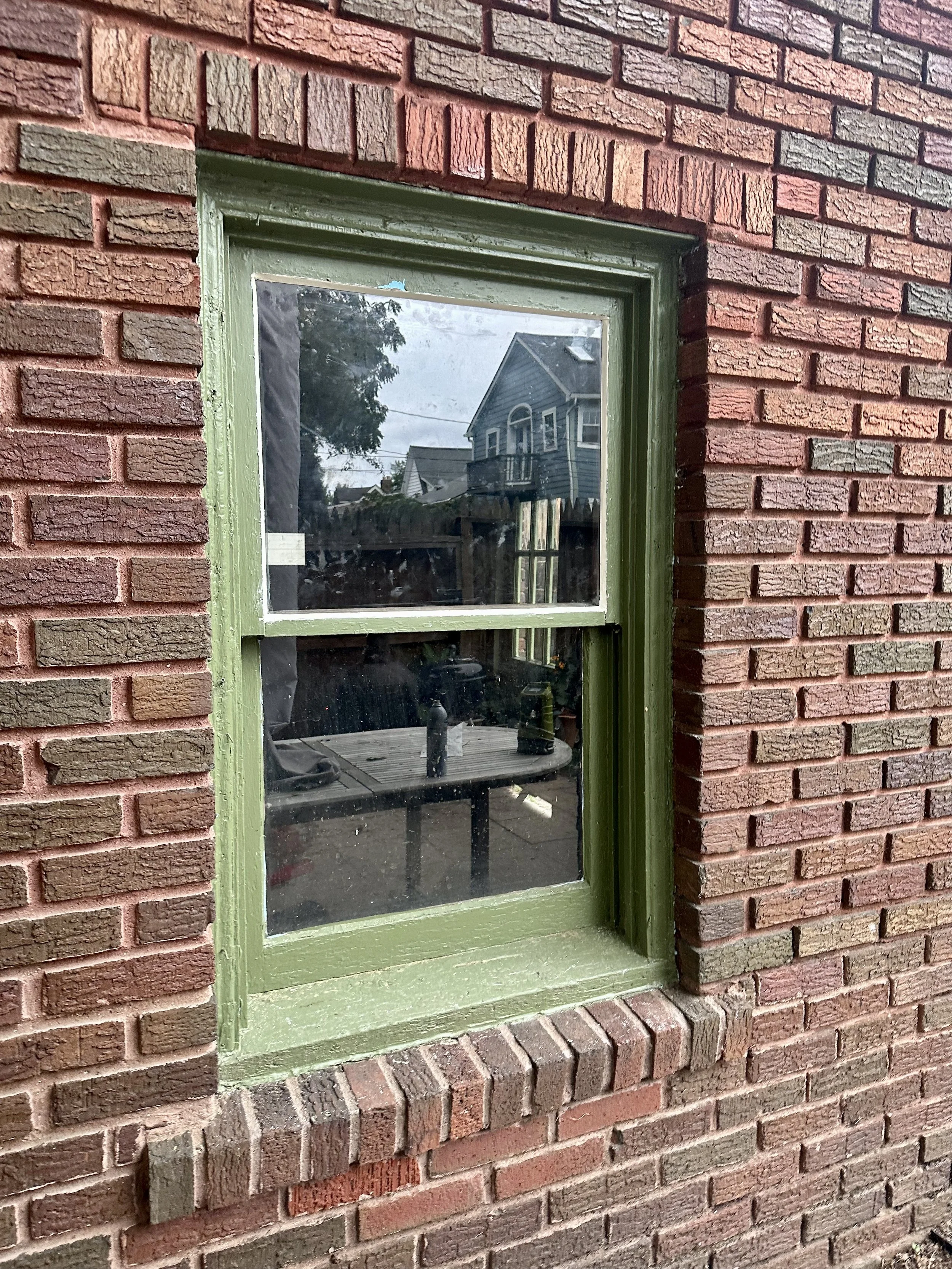 A brick house wall with a small rectangular window framed in green. The window reflects a neighboring house and trees outside.