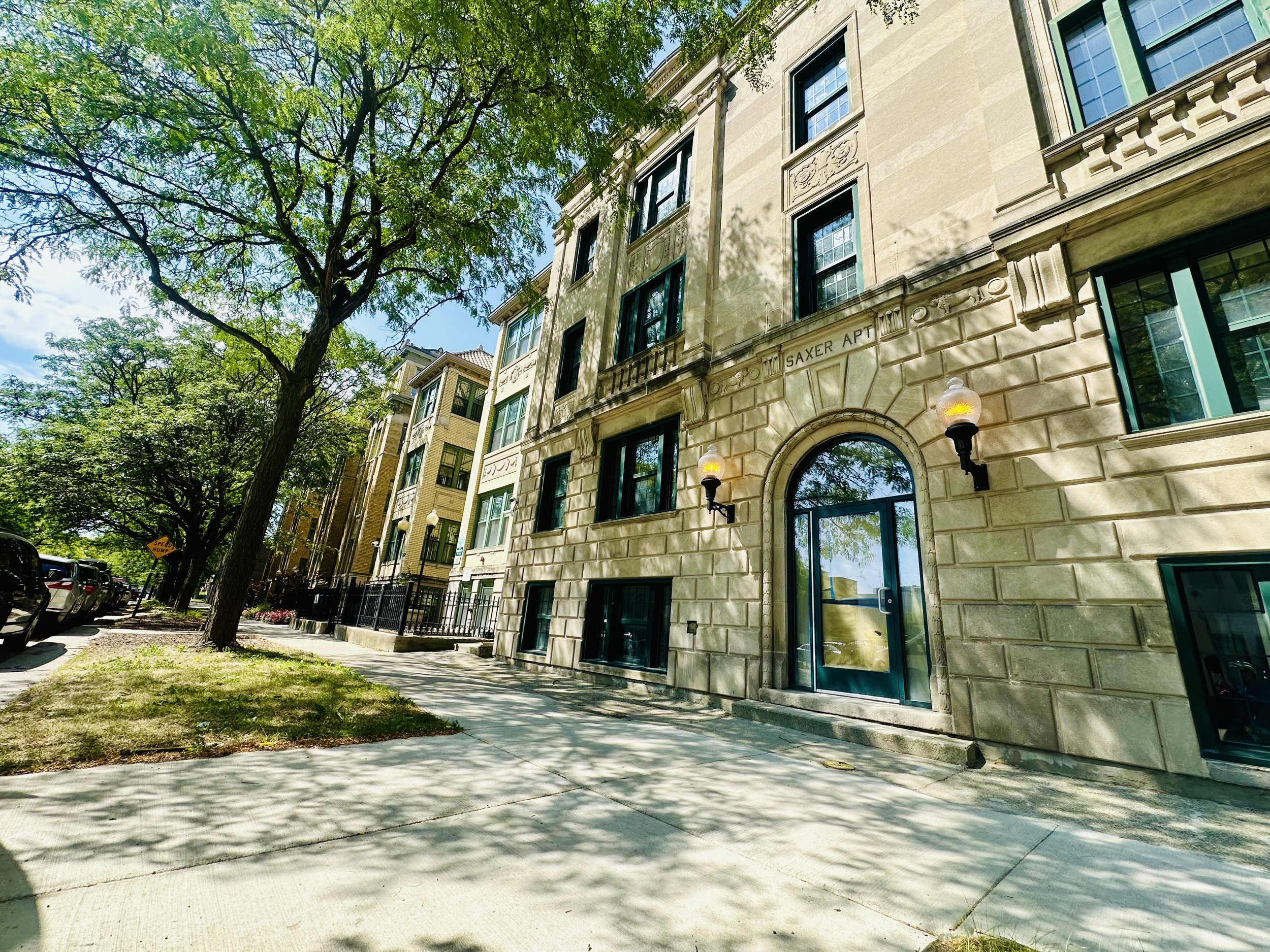 Street view of an apartment building with trees and parked cars, sunlight casting shadows on the sidewalk.