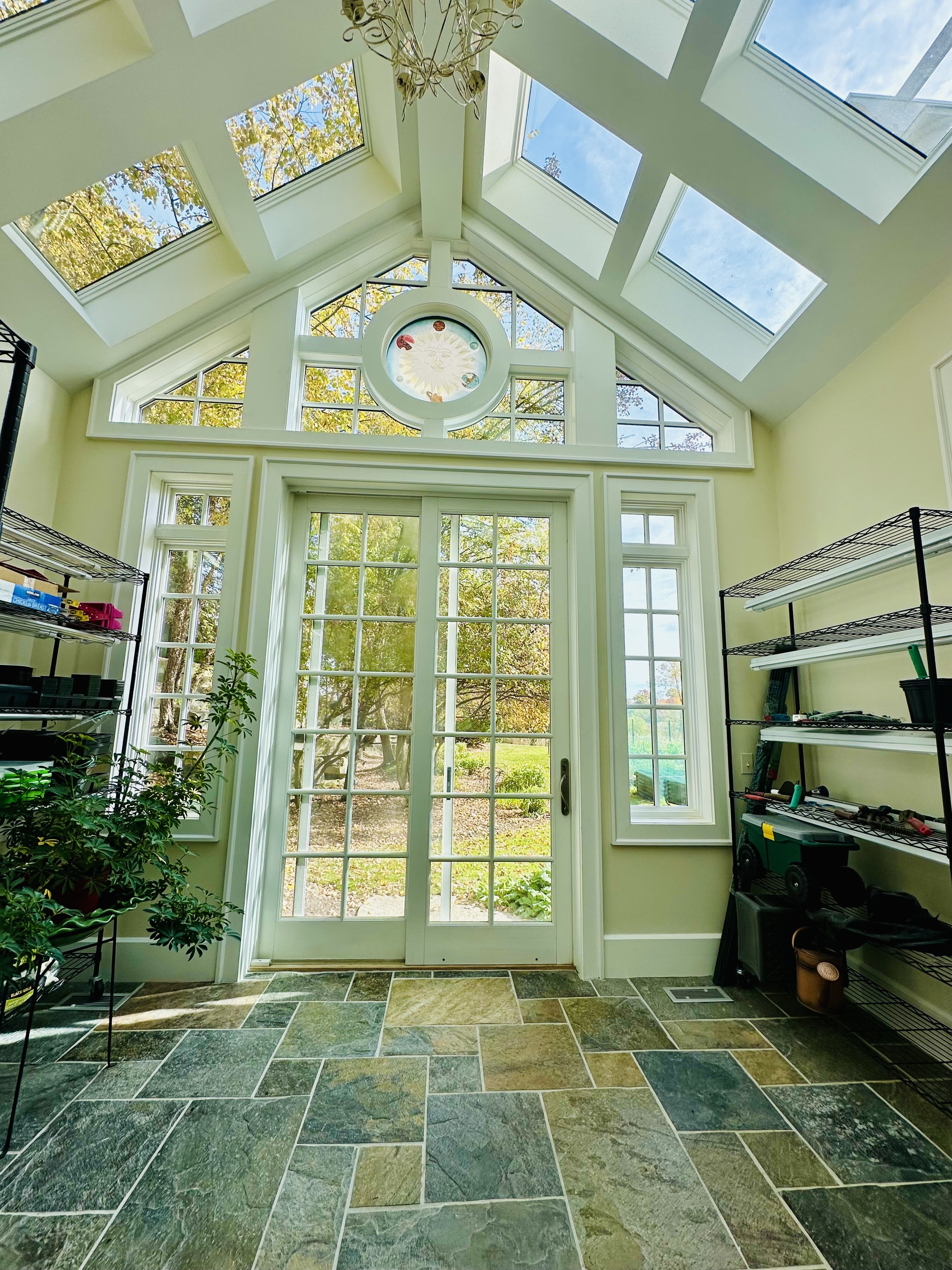 Bright sunroom with a large glass door leading outside, multiple skylights on the ceiling letting in natural light, a ceiling fan, and some shelving with gardening tools and supplies.