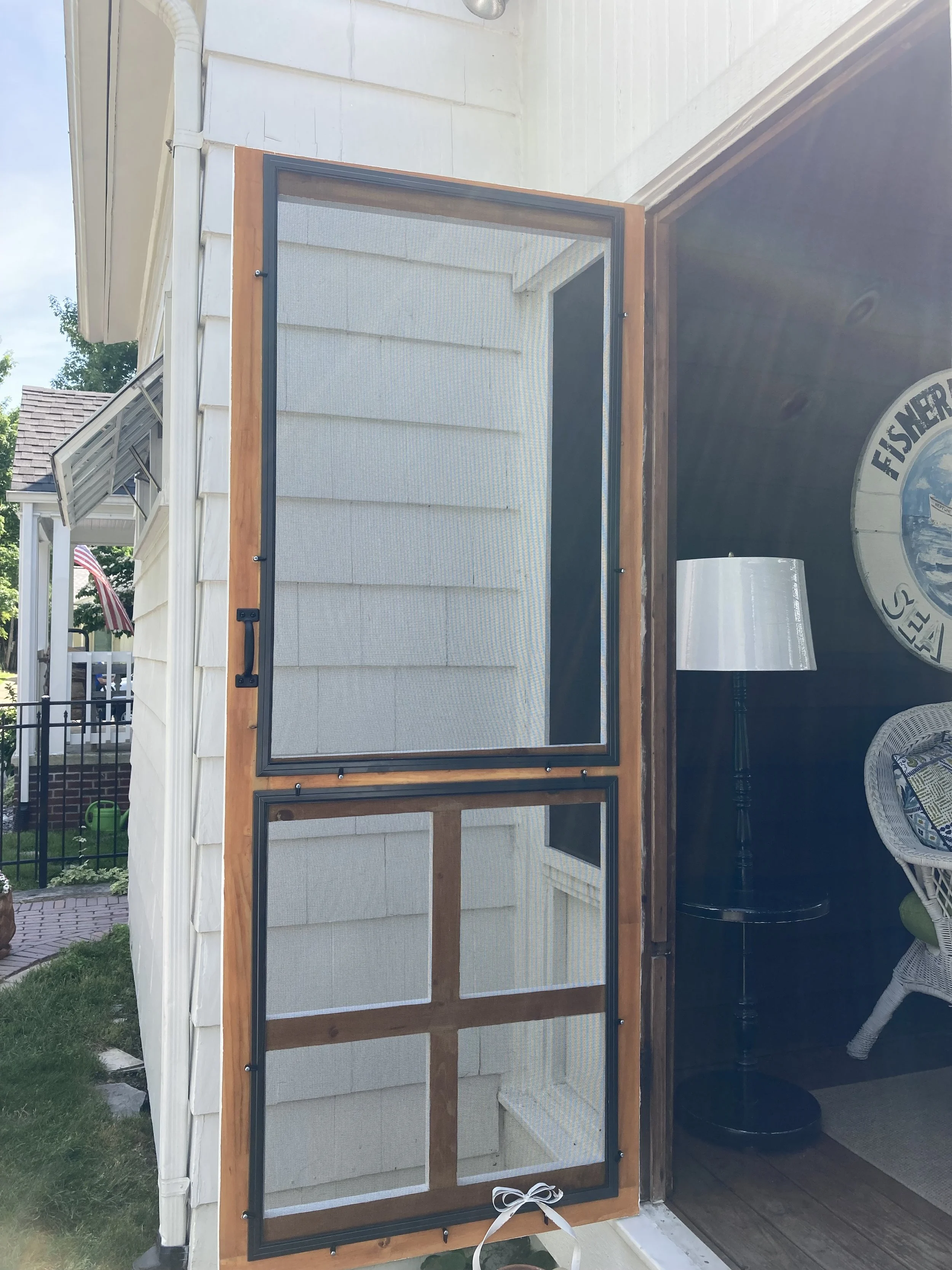 A screened wooden door with black metal frame hanging outside a house door, tied with a white ribbon at the bottom, next to a porch with a white wicker chair and a black lamp.