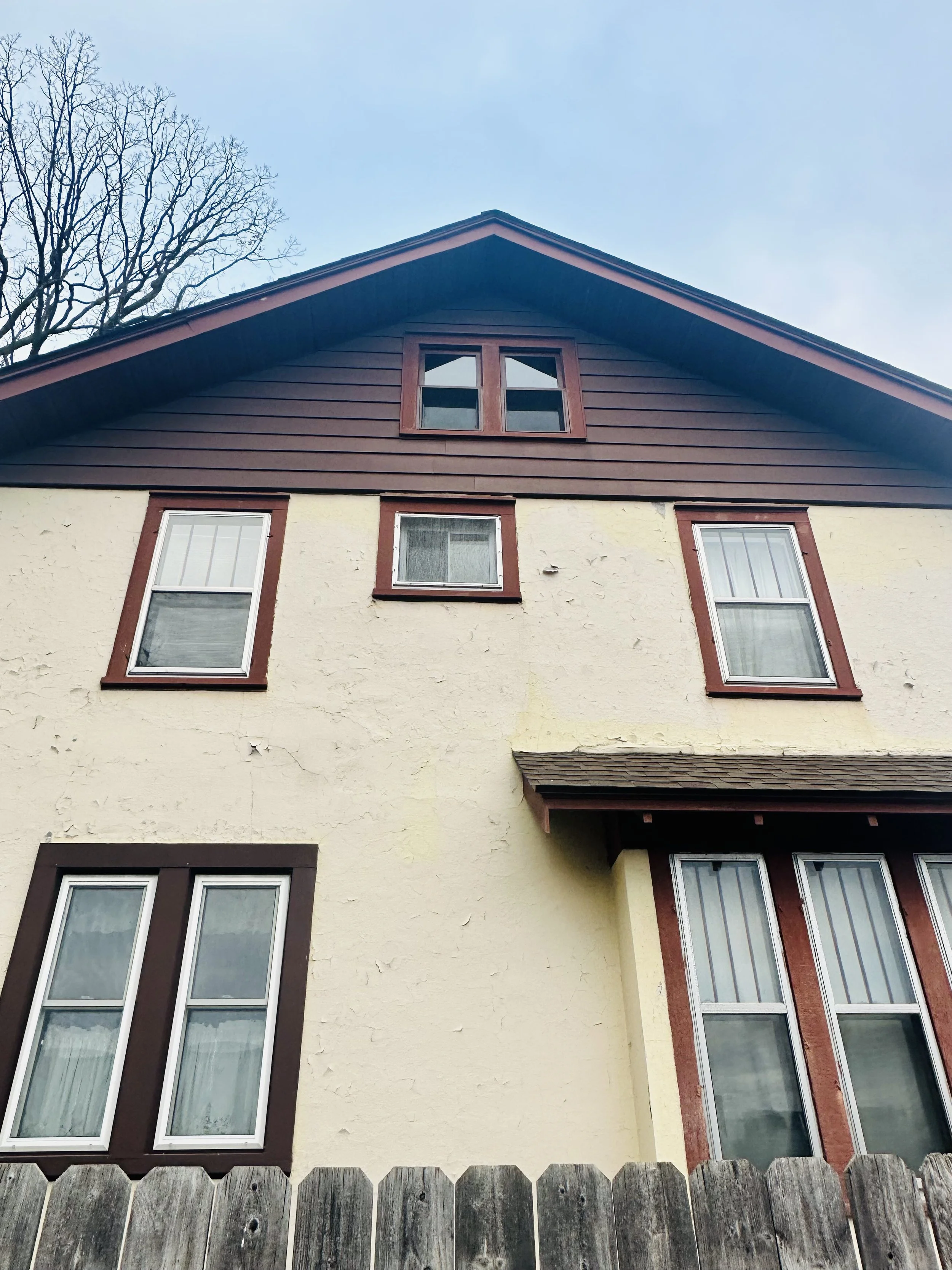 Photo of upper part of a house with cream-colored textured walls, brown trim around windows, and a steep roof with dark blue siding. There is a wooden fence in the foreground and a leafless tree in the background under a clear sky.