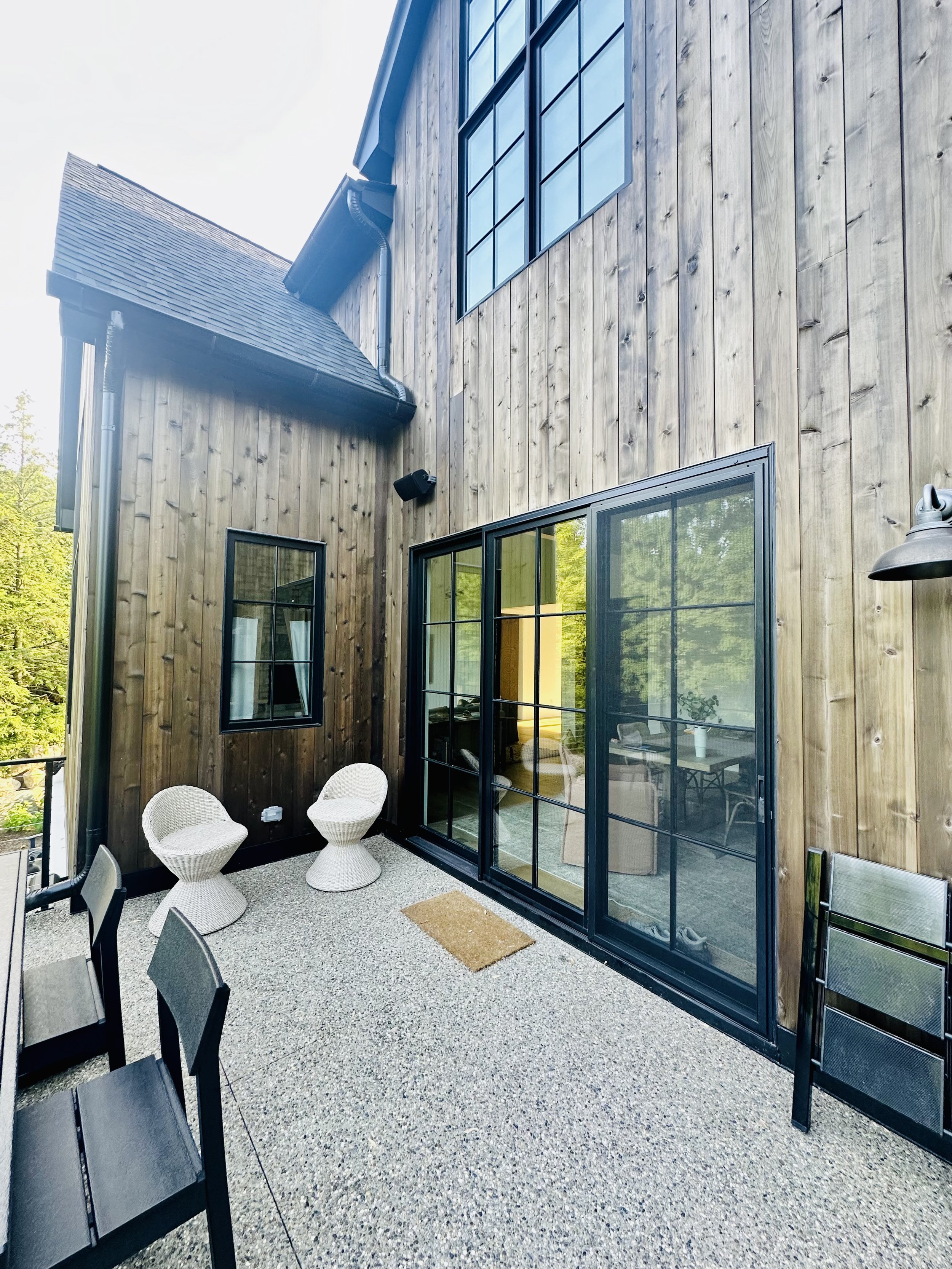 Outdoor patio with grey speckled concrete floor, black metal chairs, white woven chairs, black sliding glass door, wooden exterior wall, small window with white curtains, greenery outside, and ambient lighting.