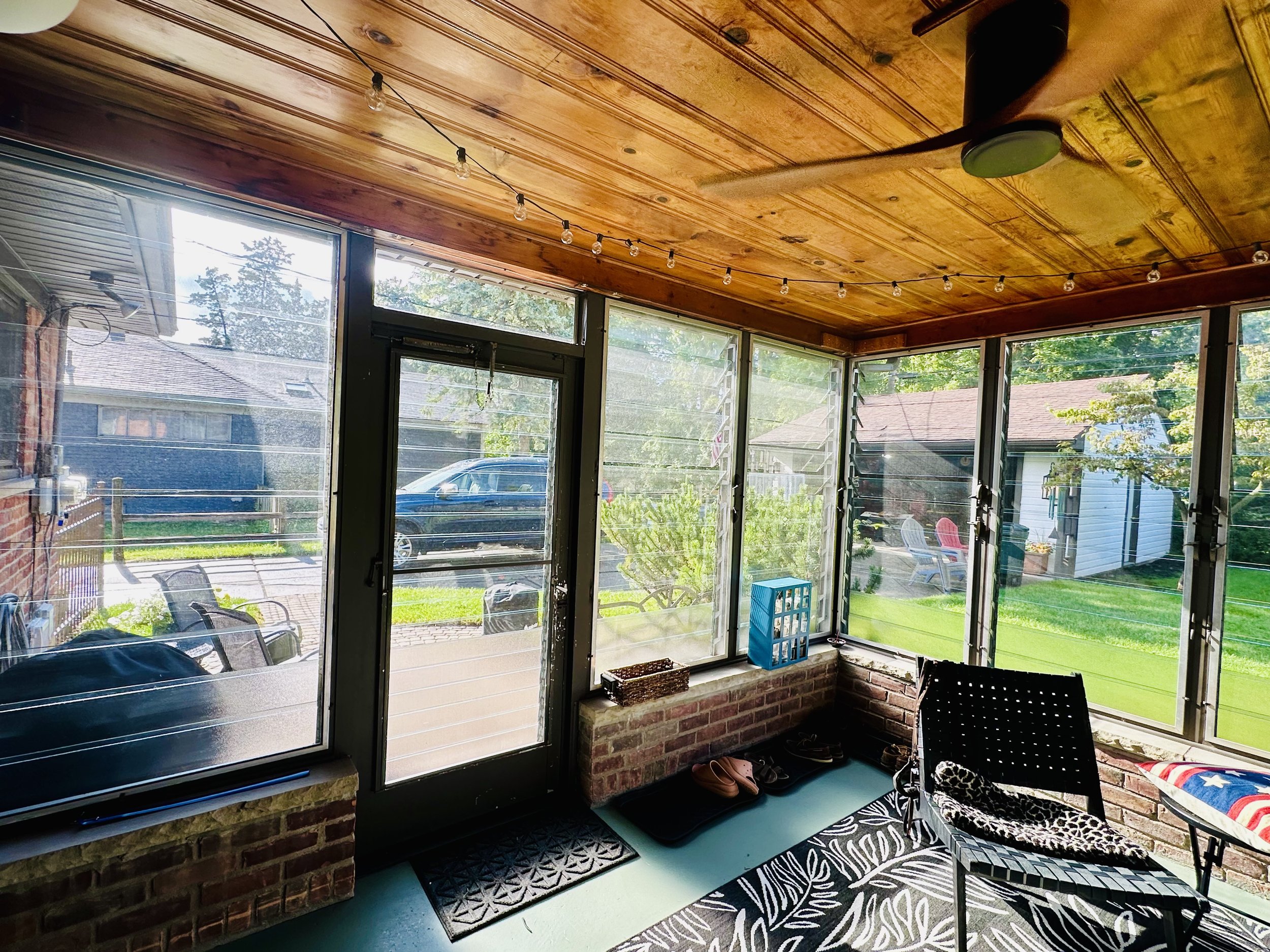 A screened porch with a wooden ceiling, string lights, and a ceiling fan. Outside, there are chairs, a shed, and a lawn with trees.