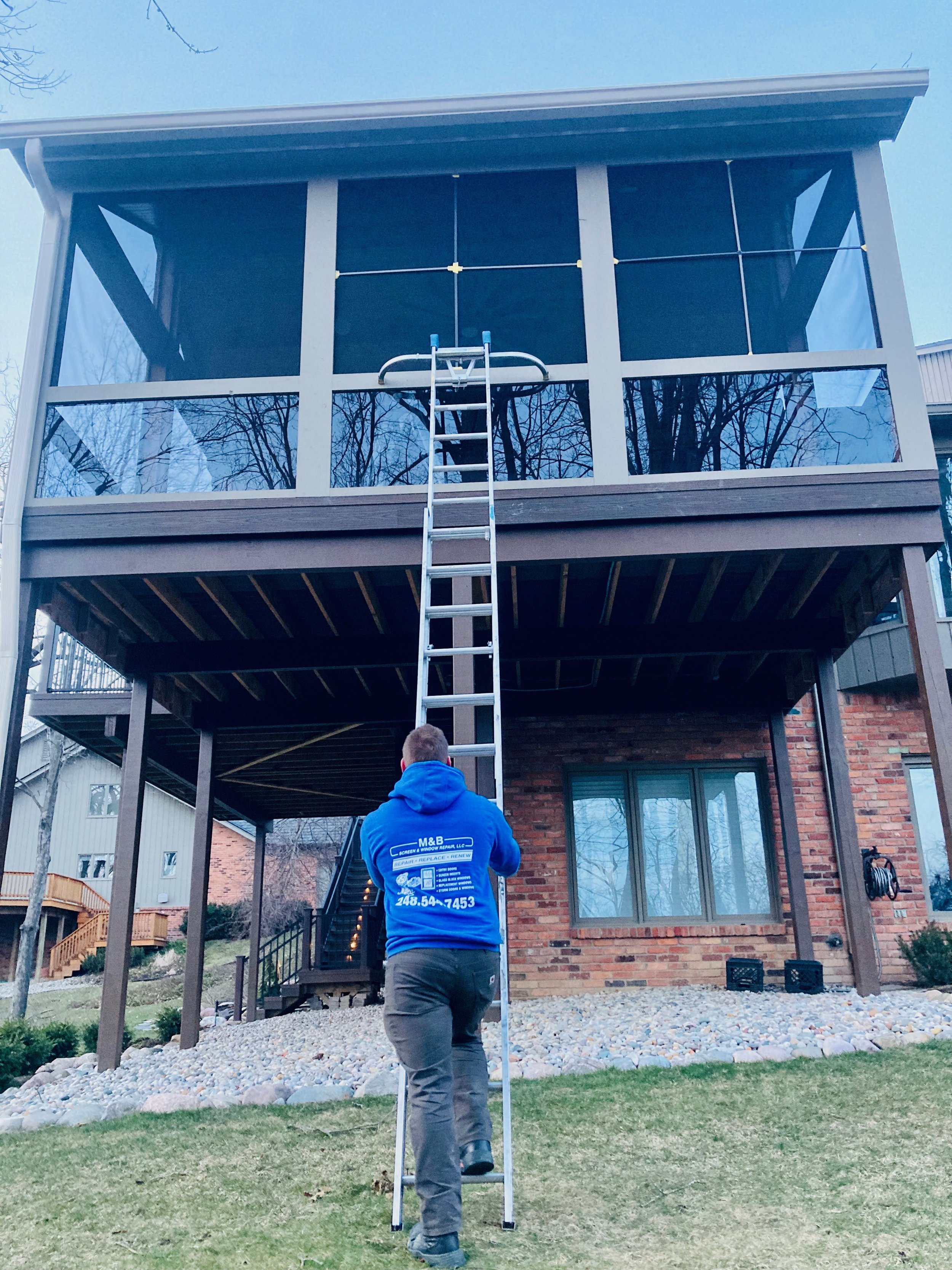 A man climbing a tall ladder towards a screened-in porch or balcony of a house. The house has a brick lower level and a wooden upper structure with large glass windows. The man is wearing a blue hoodie with some text and a phone number on the back.