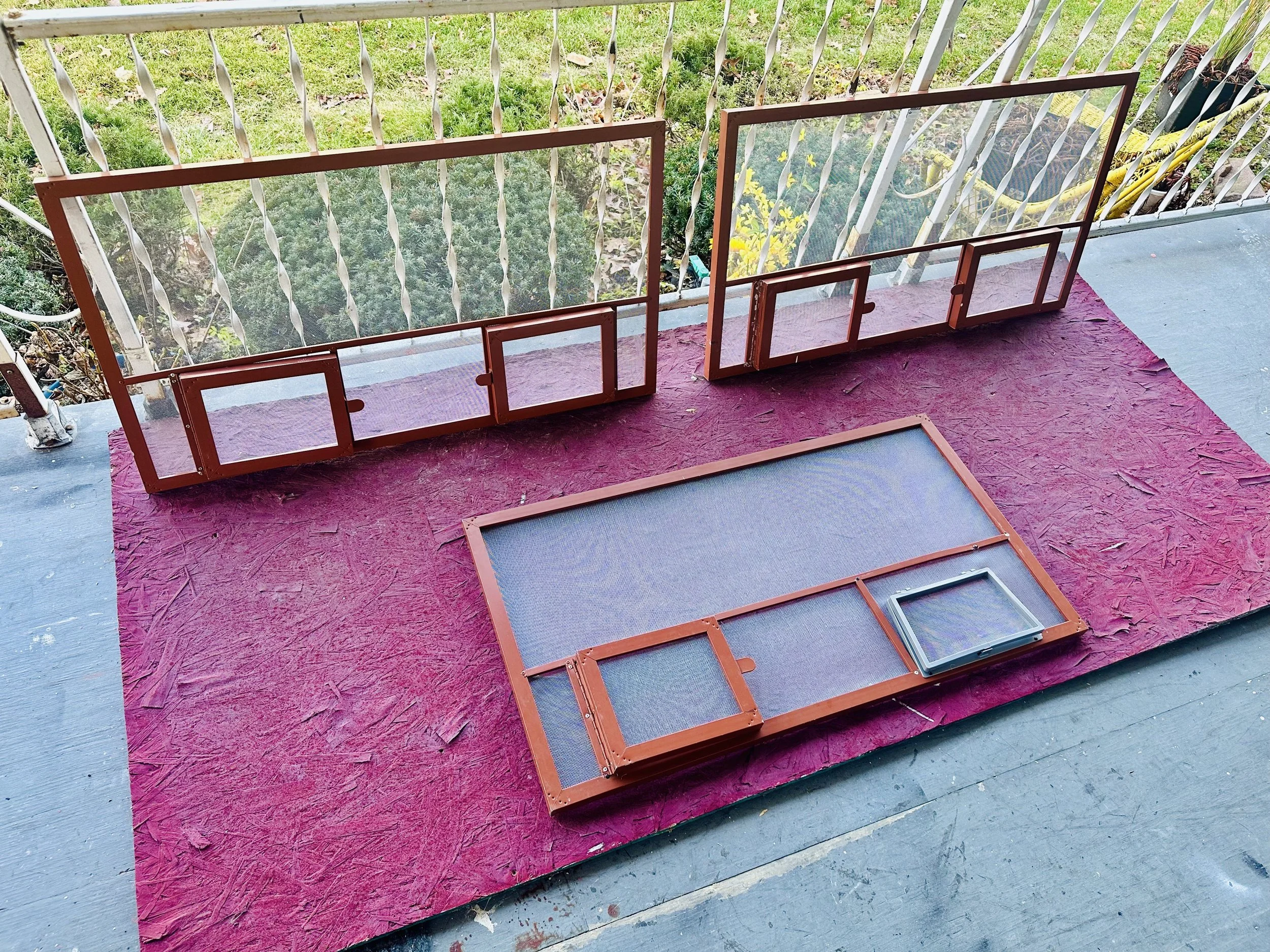Various five different sizes of window screens arranged on a pink textured outdoor carpet on a porch or balcony, with a white metal railing and some small bushes and grass visible in the background.