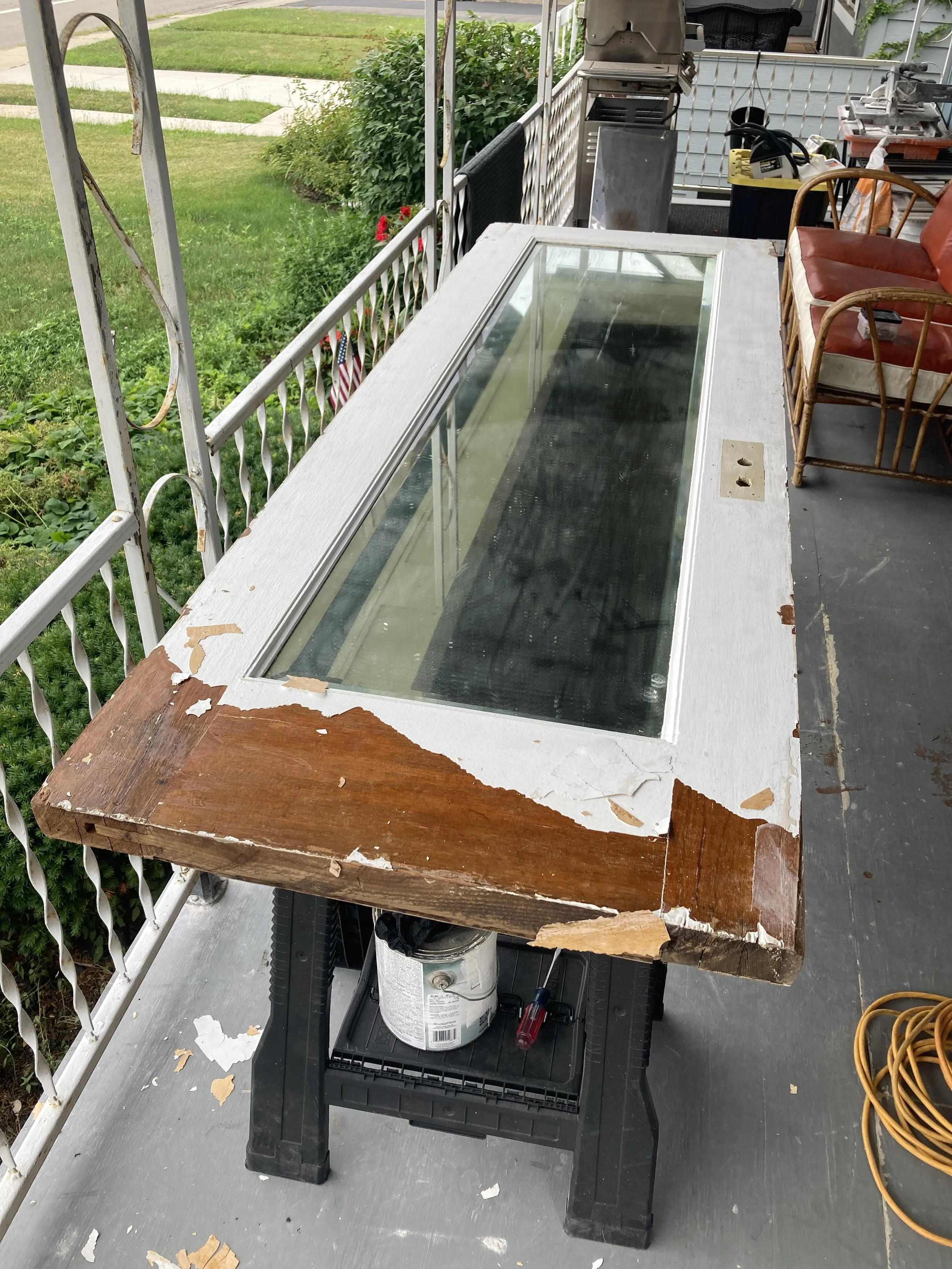 A worn wooden table with peeling white paint and glass top sitting on a black plastic step ladder, surrounded by a porch with a metal railing, chairs, and various tools and supplies.