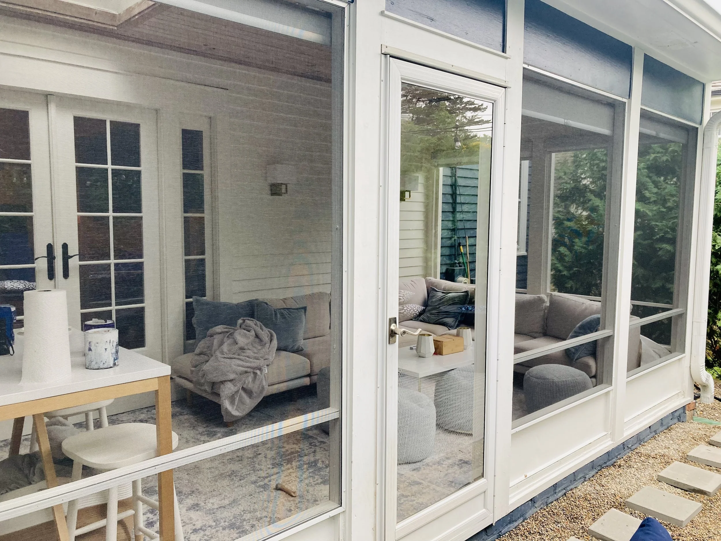 A sunroom with large glass windows and a door leading inside. Inside, there is a gray sofa with pillows, a wooden coffee table, and a dining table with chairs. Outside the sunroom, a gravel pathway and green trees are visible.