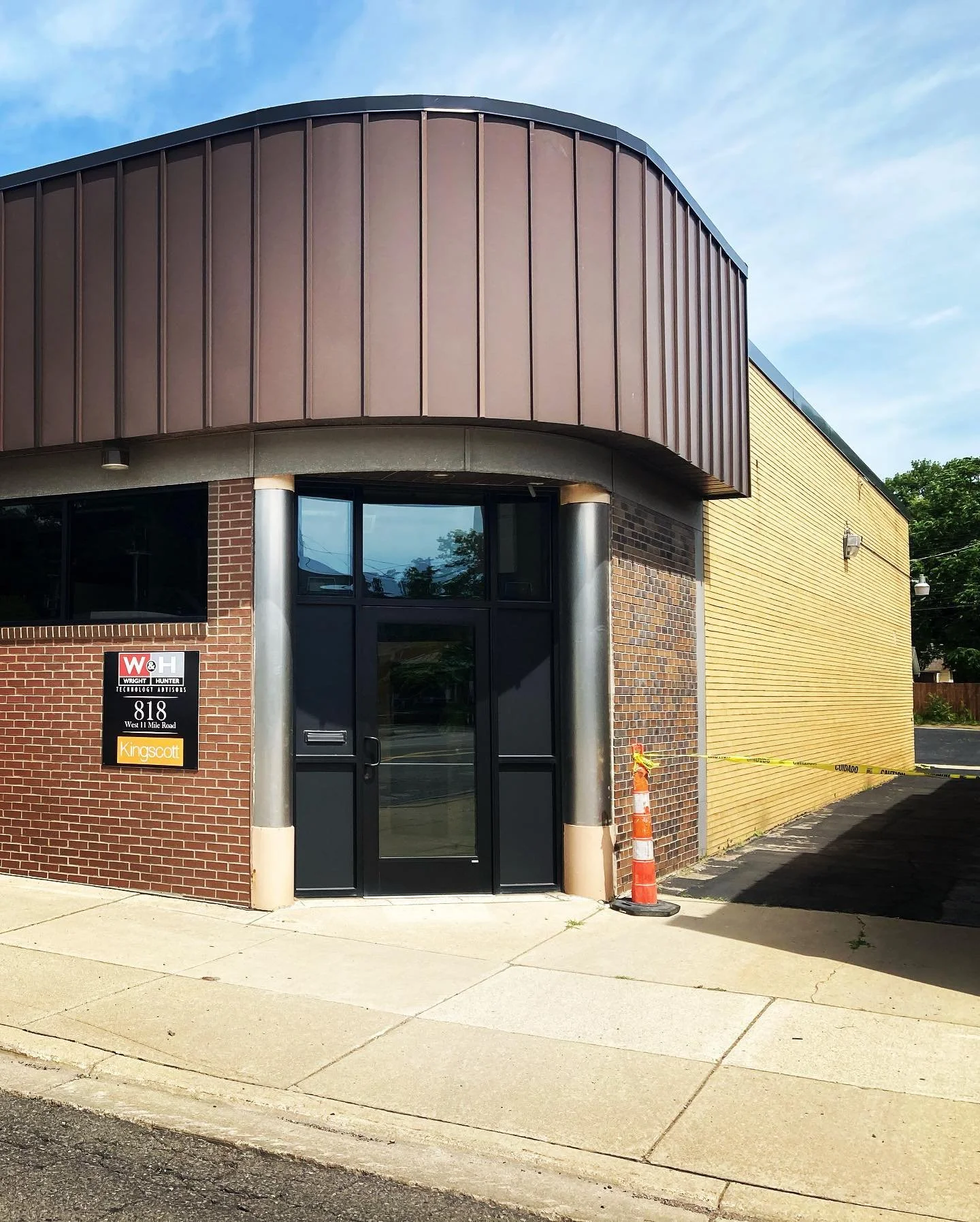 Exterior view of a commercial building with a modern design, featuring a rounded brown metal facade, brick wall, and yellow siding. There is a black door at the entrance, and a traffic cone and caution tape are on the sidewalk.