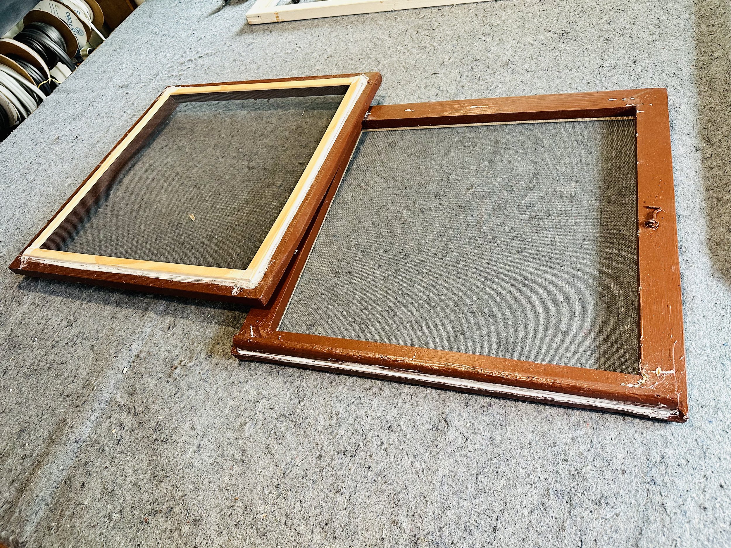 A wooden frame with glass, resting on a gray carpeted surface, with part of a workshop or store environment visible in the background.