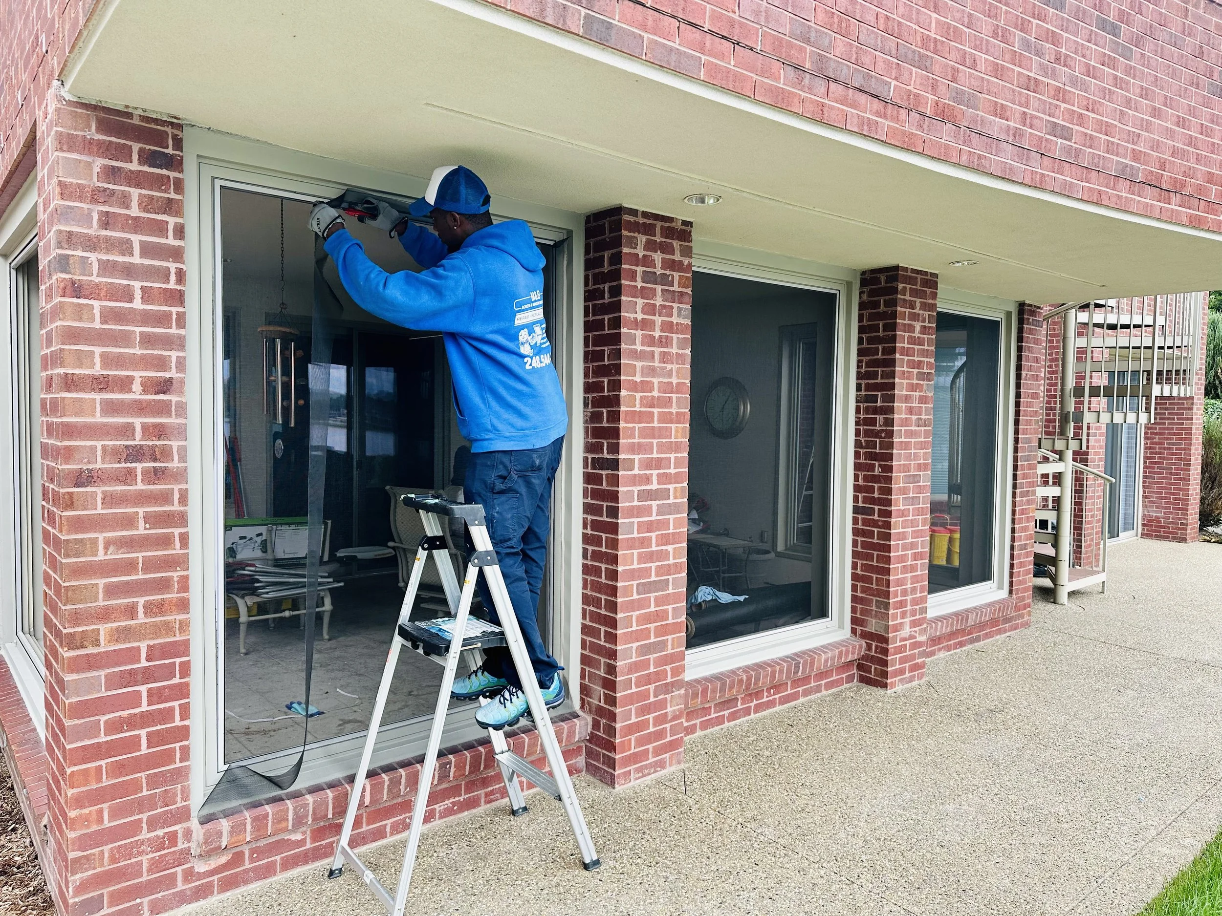 A man standing on a step ladder working on the top of a large glass sliding door in a brick house.
