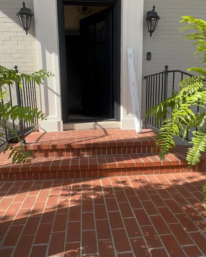 Brick steps leading up to a dark front door with black frame, flanked by black wall-mounted lanterns and a white wall. A white door frame is visible, with a white sign or board leaning against the wall to the right. Green plants are on both sides of