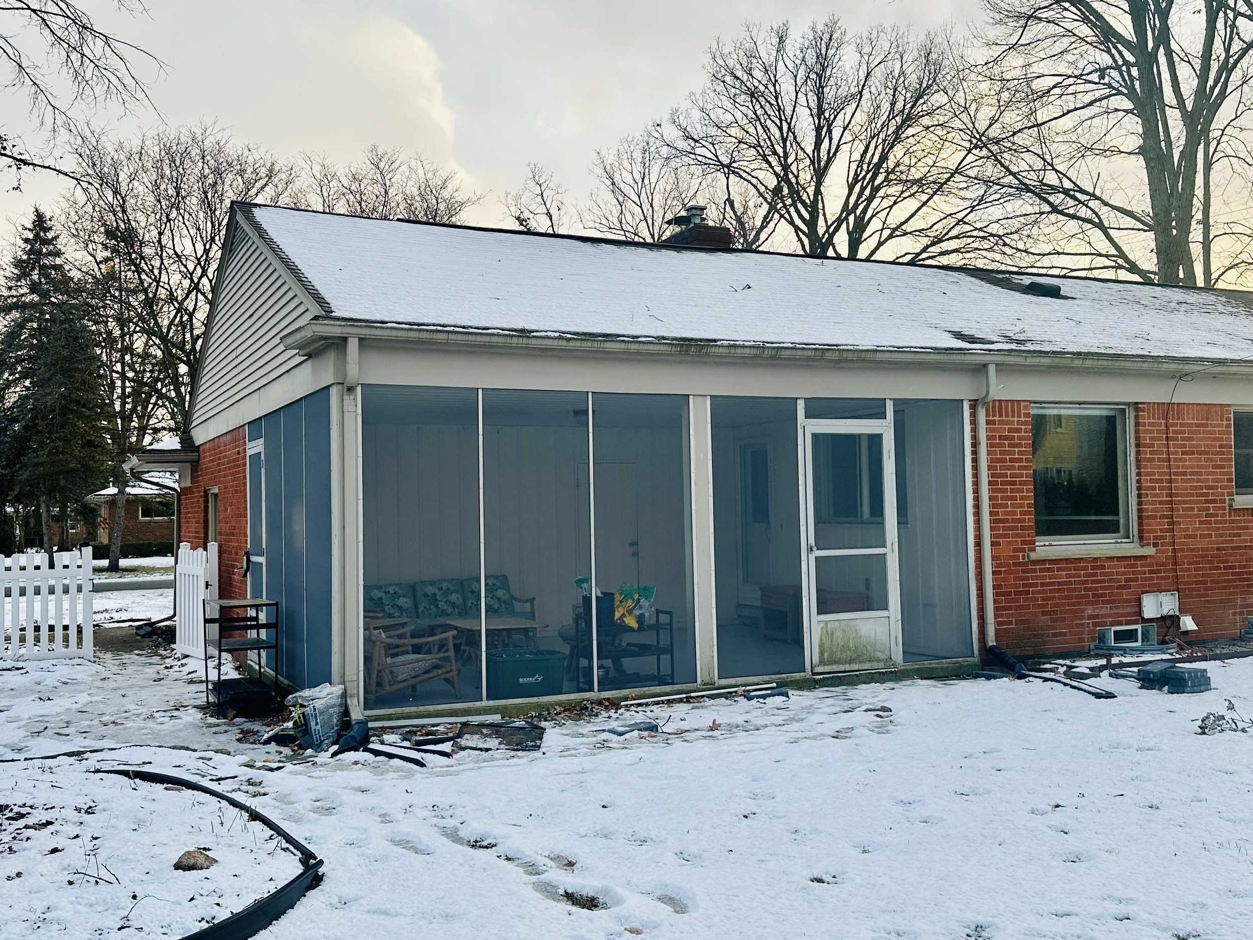 Backyard with snow-covered ground and a house with a porch and large windows. The house has a combination of brick and siding exterior. The roof is also covered in snow. There are some scattered debris and gardening tools in the yard, and leafless tr