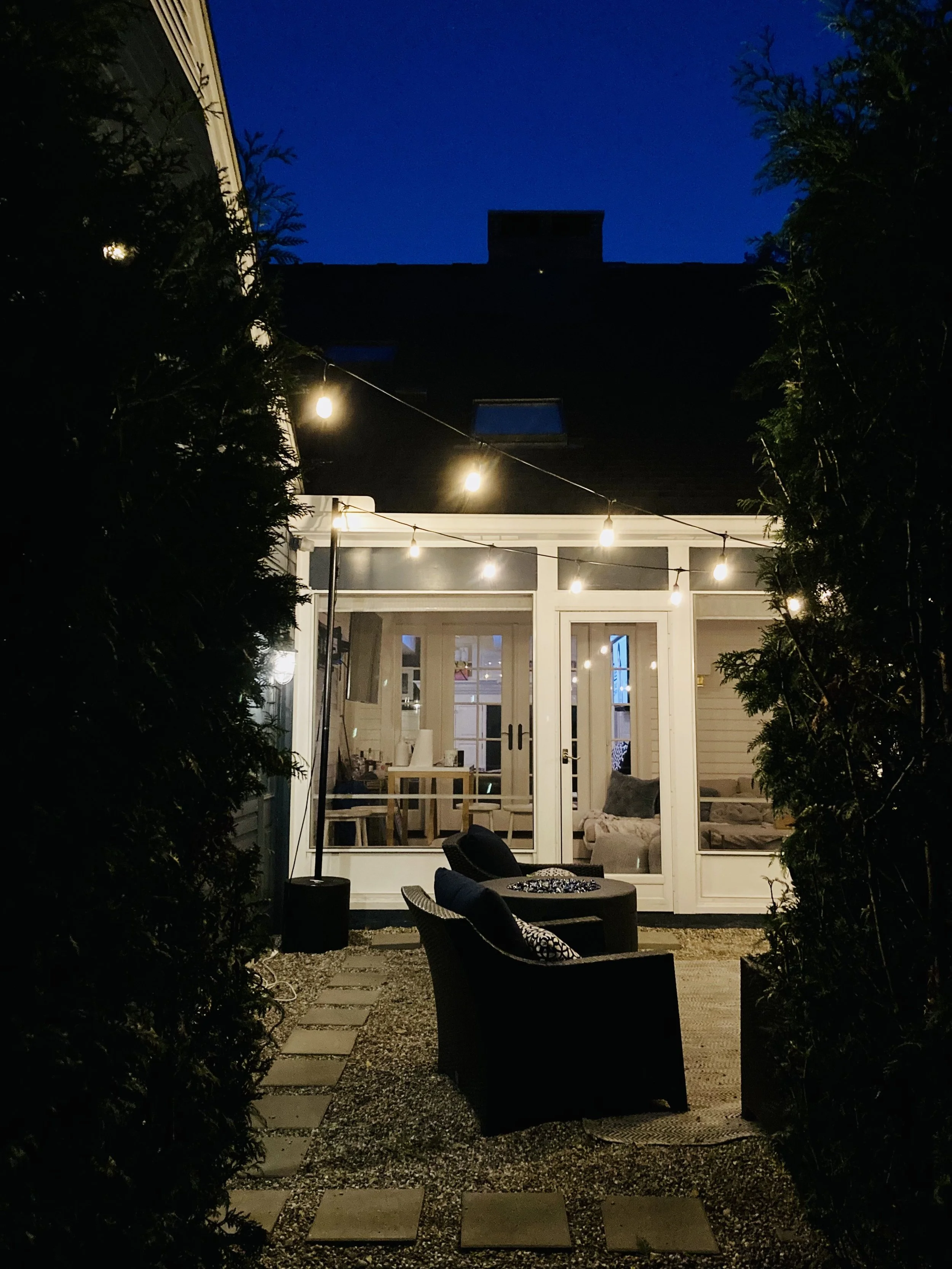 Nighttime view of a backyard patio with string lights, outdoor chairs, and a house with large glass doors leading inside, surrounded by trees.