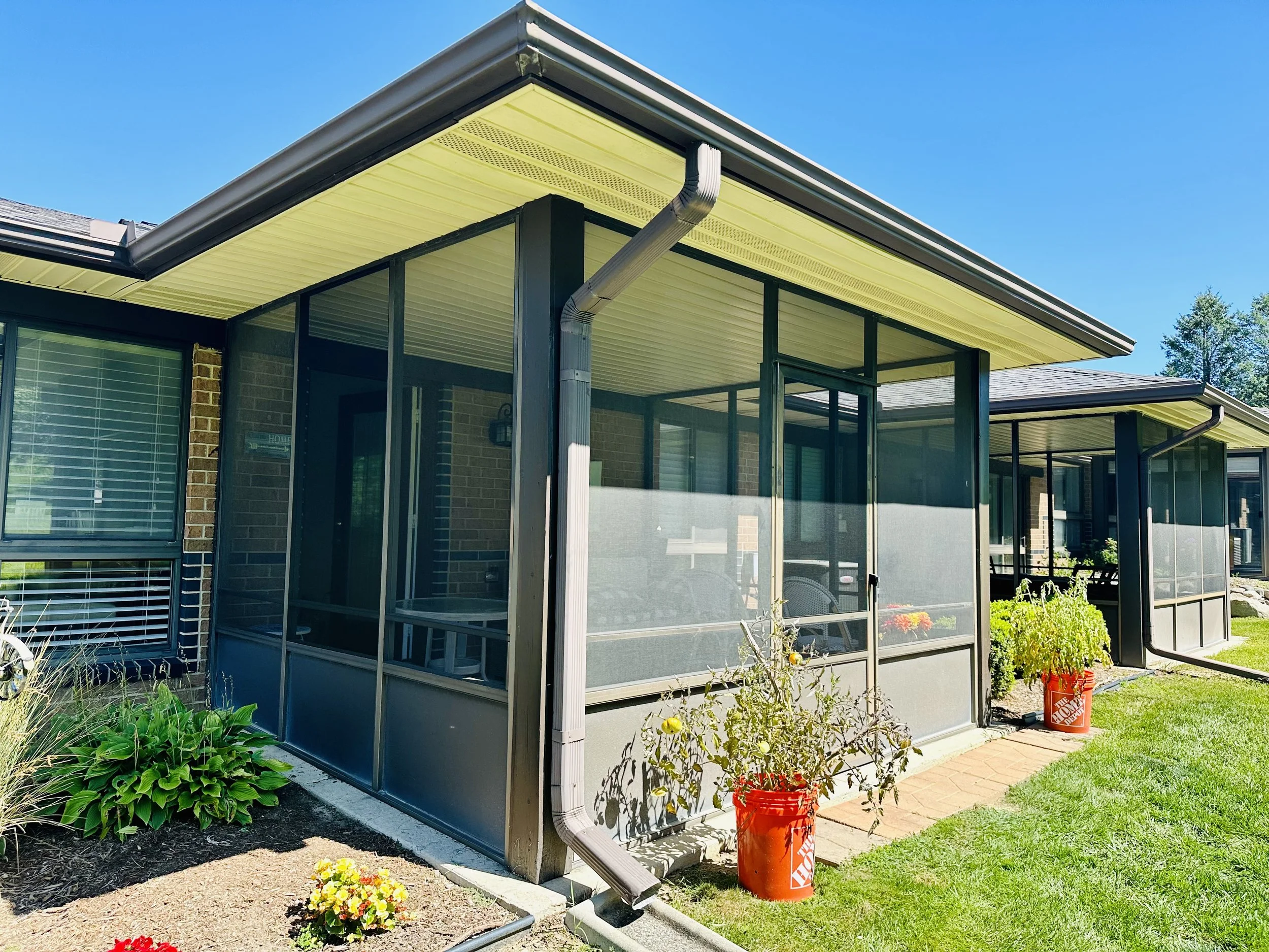 A screened porch on a brick house with gardening plants and red flower pots outside under a clear blue sky.