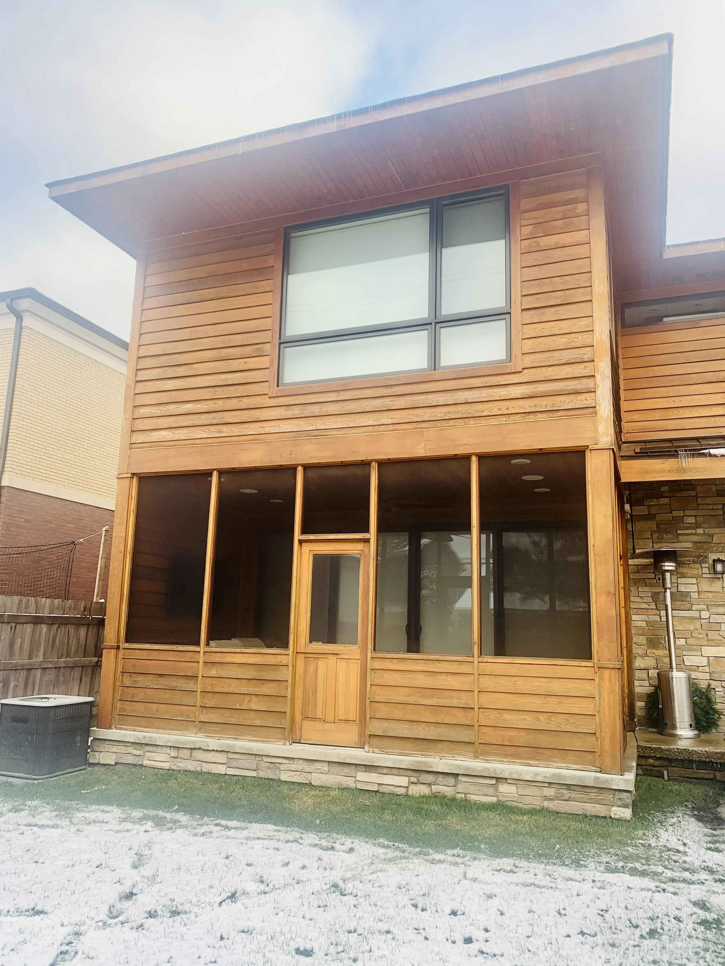 Front view of a two-story house with a wooden frame, large windows, and a partial screened porch, with snow on the ground.