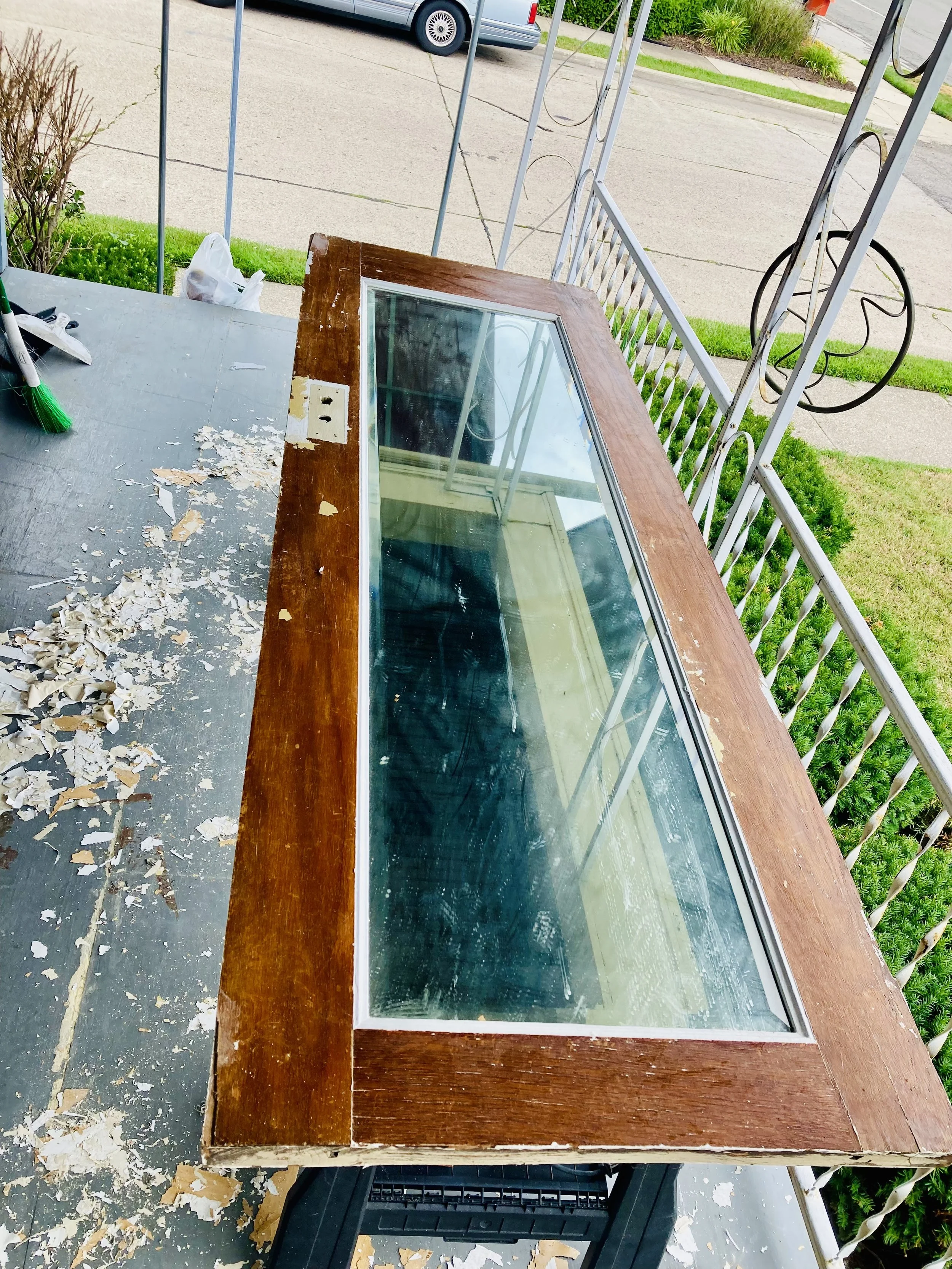 A vintage wooden and glass display case or cabinet on a porch, next to a patio railing. The wood appears worn, and the glass is in good condition.