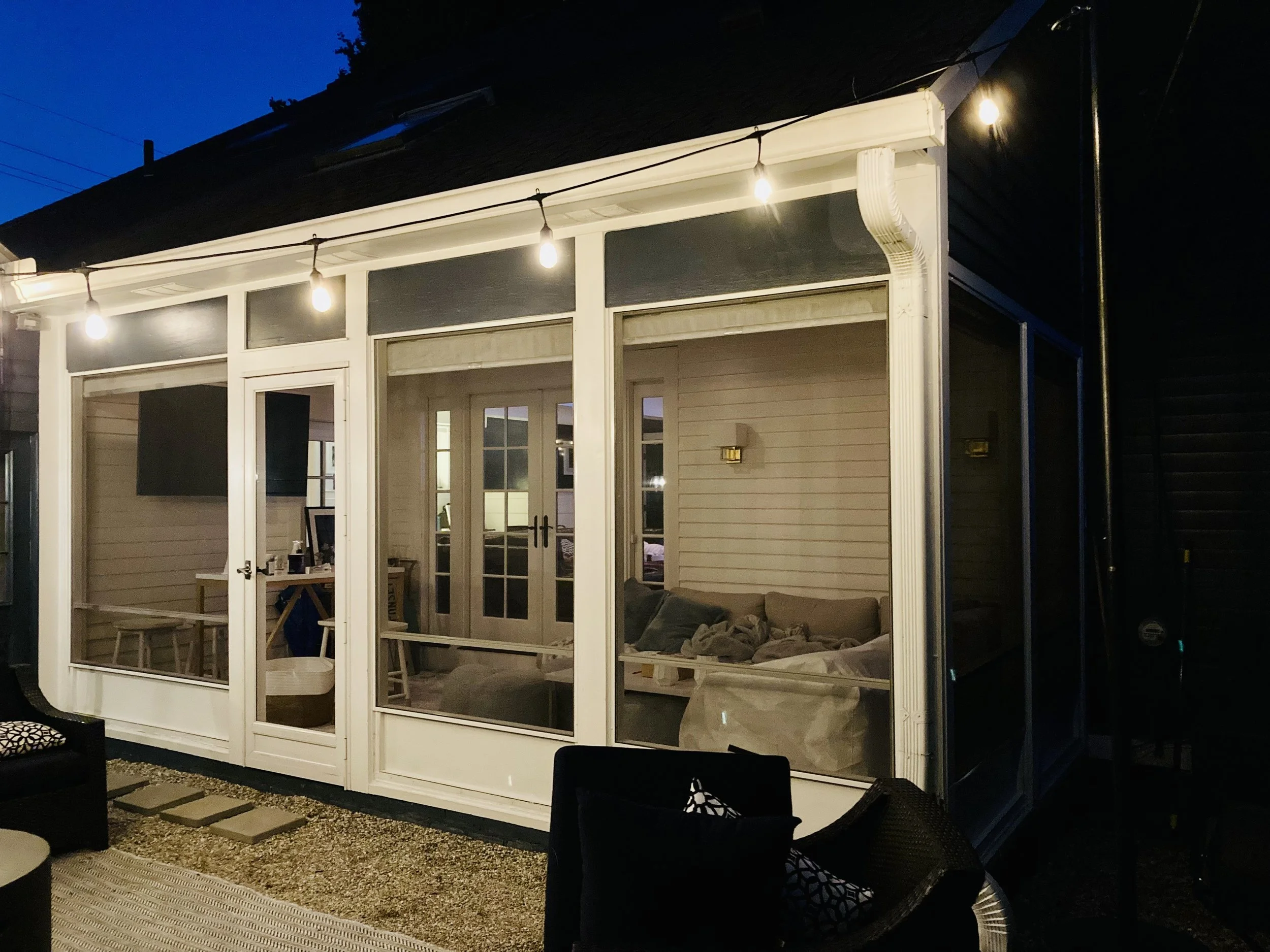 A house with a porch enclosed by white framing and large glass windows and doors. String lights hang across the porch exterior. Inside, a sofa and a table are visible through the windows, with some furniture and decor seen inside the house. The scene