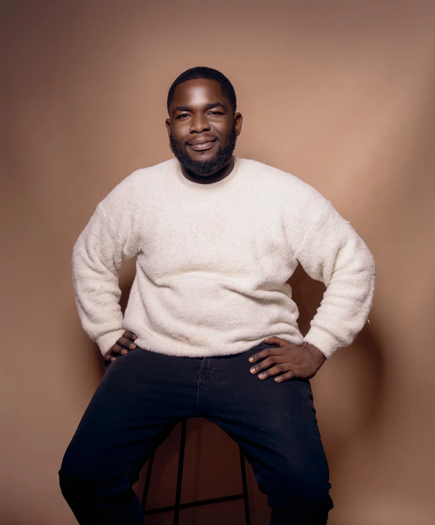 A man with dark skin, short hair, and a beard, sitting on a stool with a tan background, wearing a white fluffy sweater and black pants.