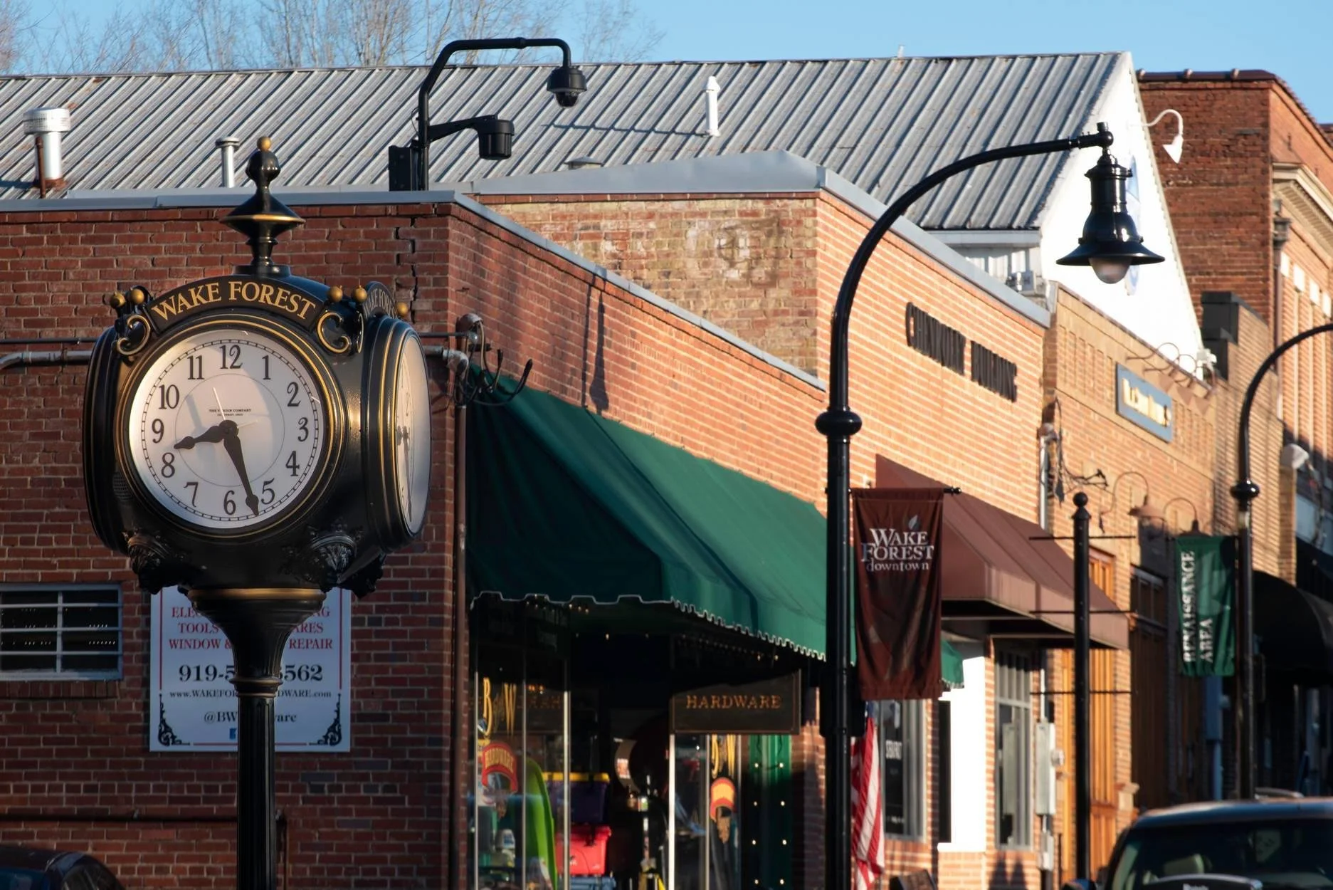 Clock on a pole reading 5:26 in front of a brick building with store signs and street lamps.