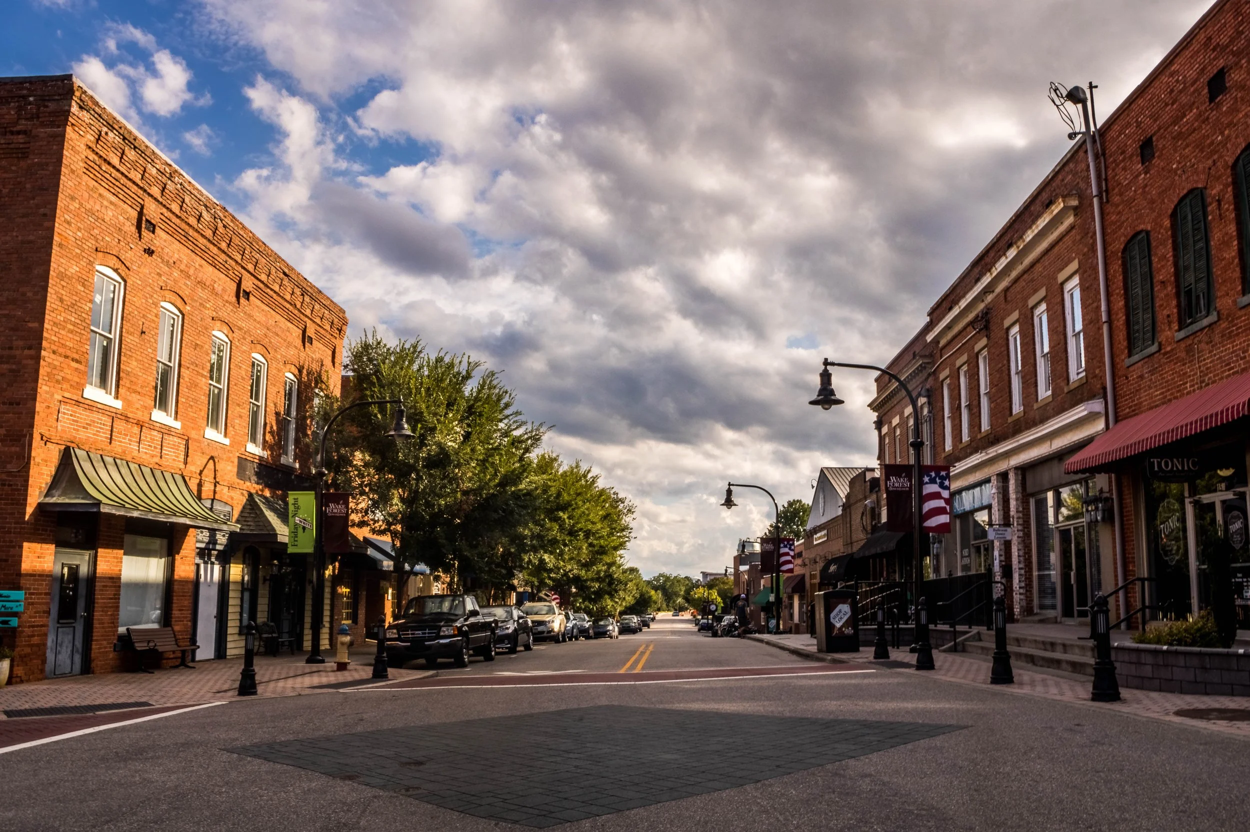 A small town street with brick buildings, parked cars, street lamps, and trees, under a partly cloudy sky.