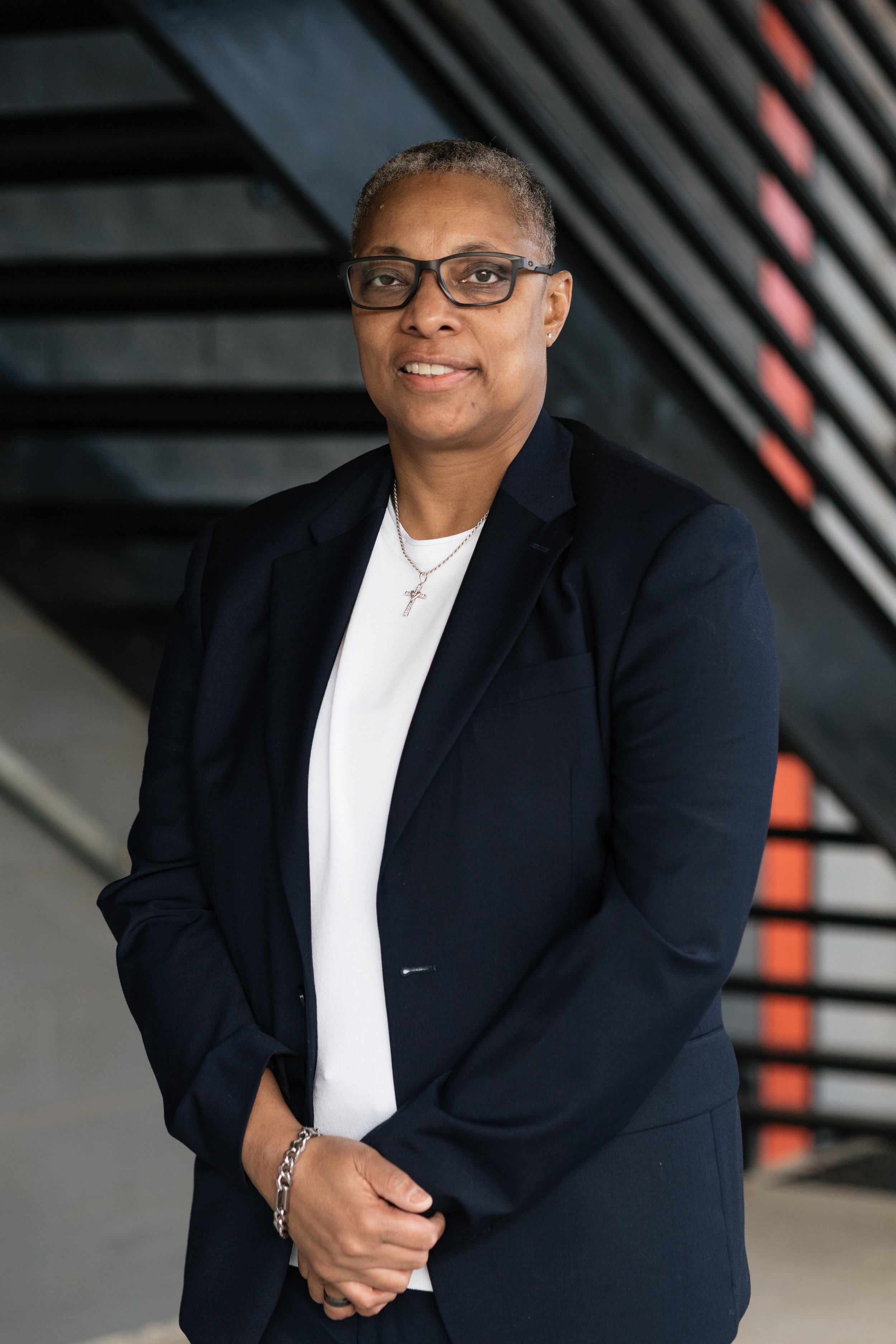 A woman with short gray hair, glasses, and earrings wearing a navy blazer, white shirt, and necklace with a cross pendant, standing indoors against a staircase with black steps and orange accents.
