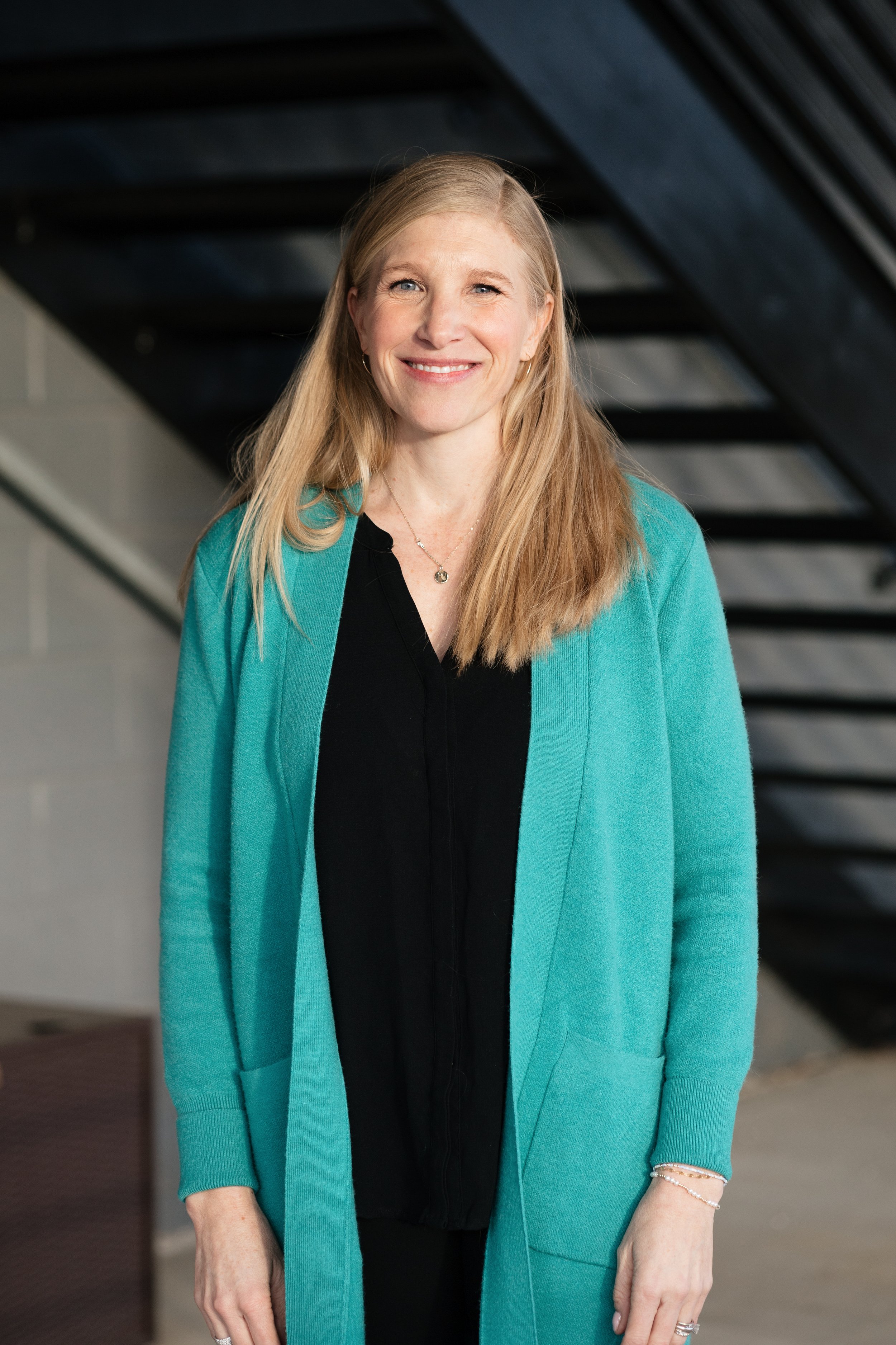 A woman with long blonde hair smiling, wearing a black top, turquoise cardigan, and jewelry, standing indoors with a dark staircase in the background.
