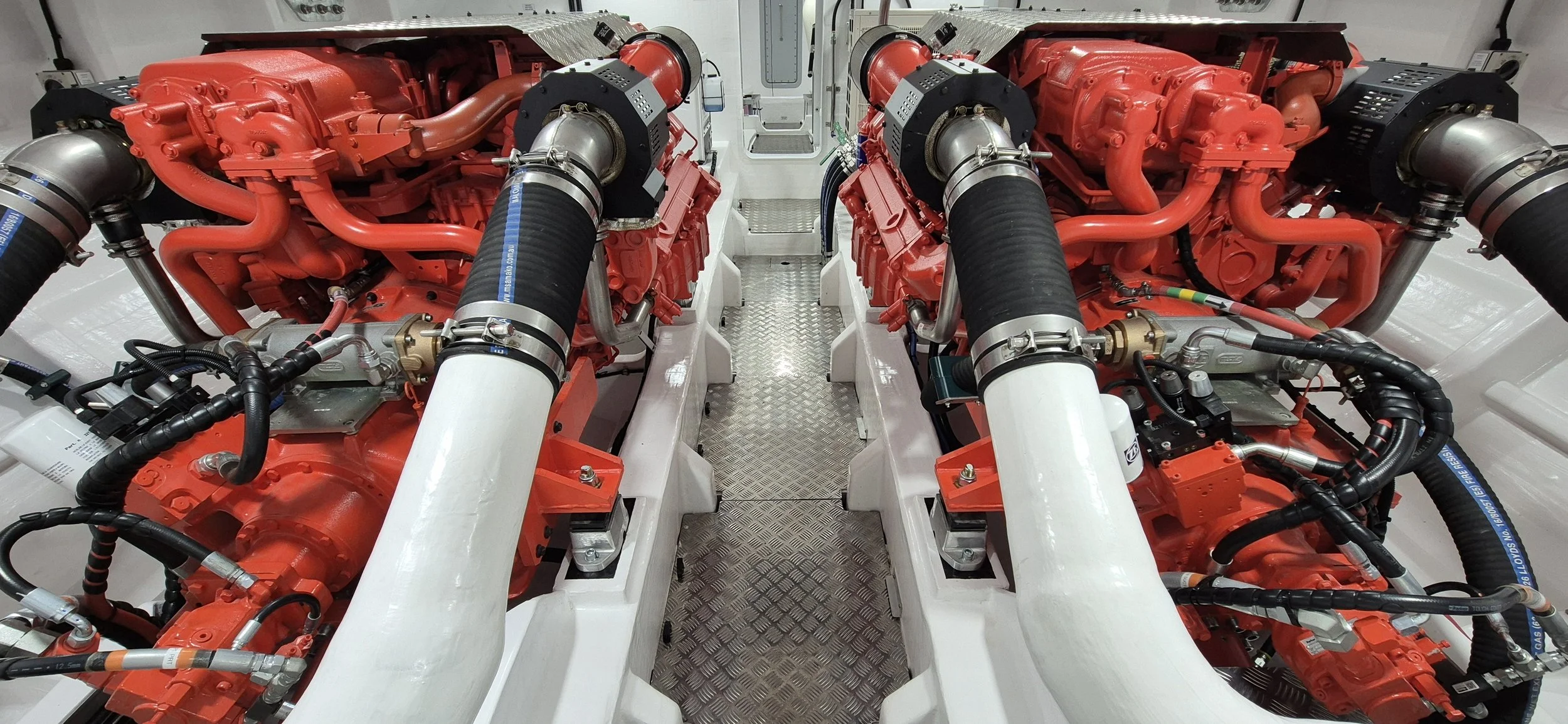 Interior of a boat engine room with two large diesel engines, metal railings, and various mechanical components, such as filters and hoses.