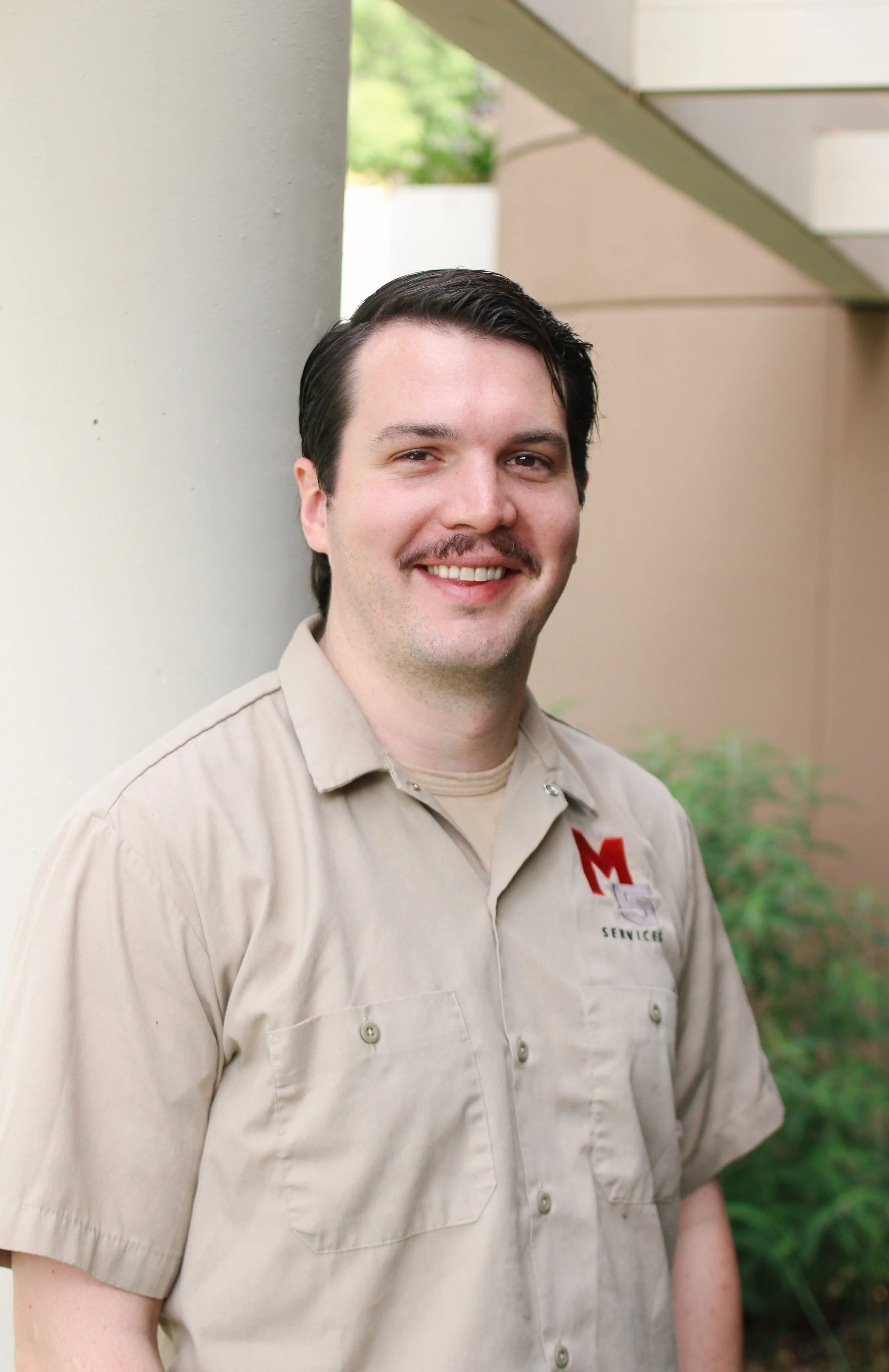 A man with dark hair, mustache, and light skin smiling outdoors, wearing a blue polo shirt with a red and white logo that says 'M5 Services'.