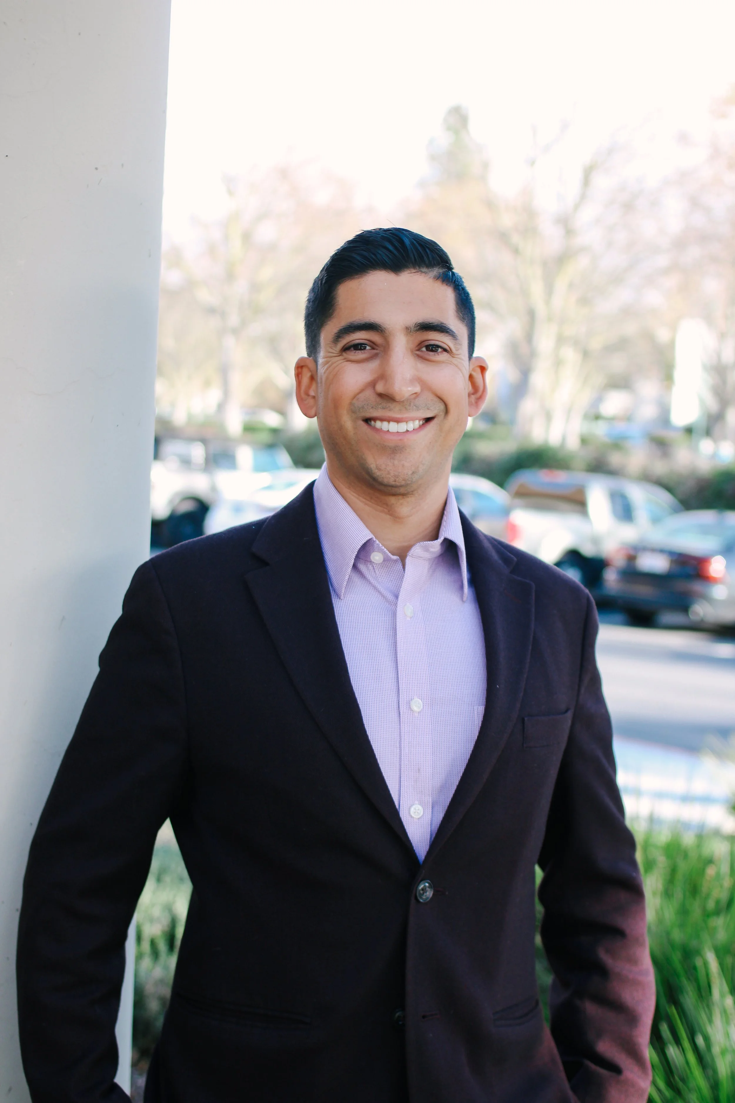 A young man and a bright smile, wearing a navy blazer with gold buttons and a gray top, standing outdoors with trees and modern buildings in the background.